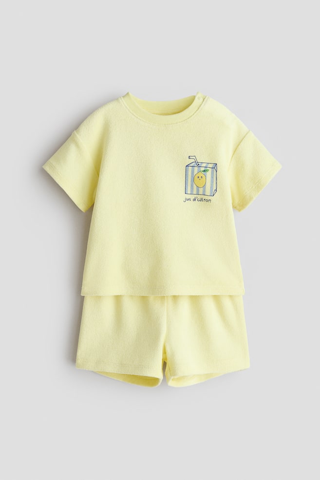 Un ensemble bébé deux pièces jaune est présenté de face, comprenant un haut en tissu éponge à manches courtes et un short assorti. Le haut présente un col rond avec des boutons-pression à l'épaule gauche et un graphique d'une boîte de jus rayée avec un visage de citron et le texte "jus d'citron" sur la poitrine. - 1