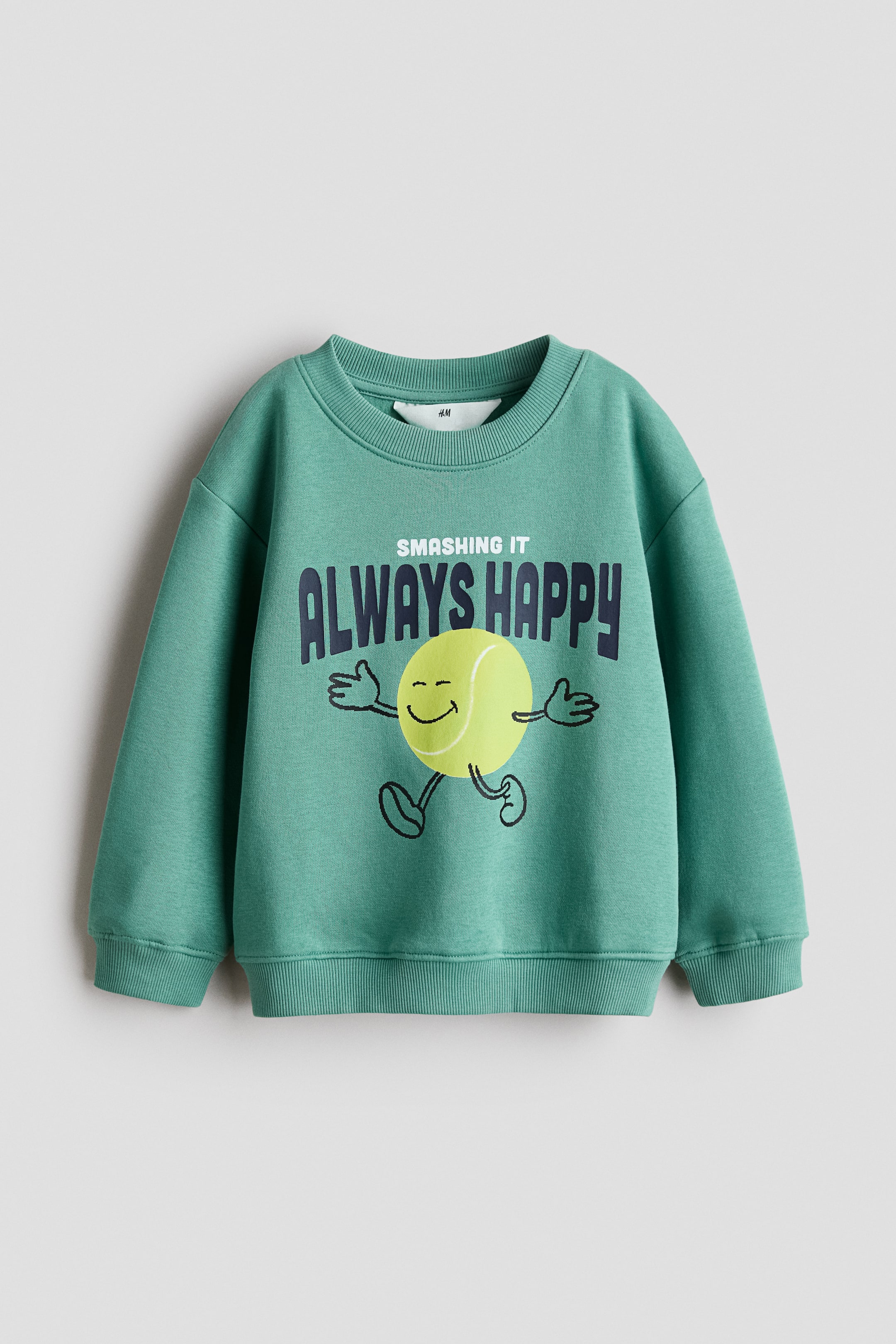 View larger image: A green jumper featuring a ribbed crew neck, long sleeves with ribbed cuffs, and a ribbed hem. The front of the jumper displays a graphic print with the text "SMASHING IT ALWAYS HAPPY" and a smiling cartoon tennis ball character with arms and legs.
