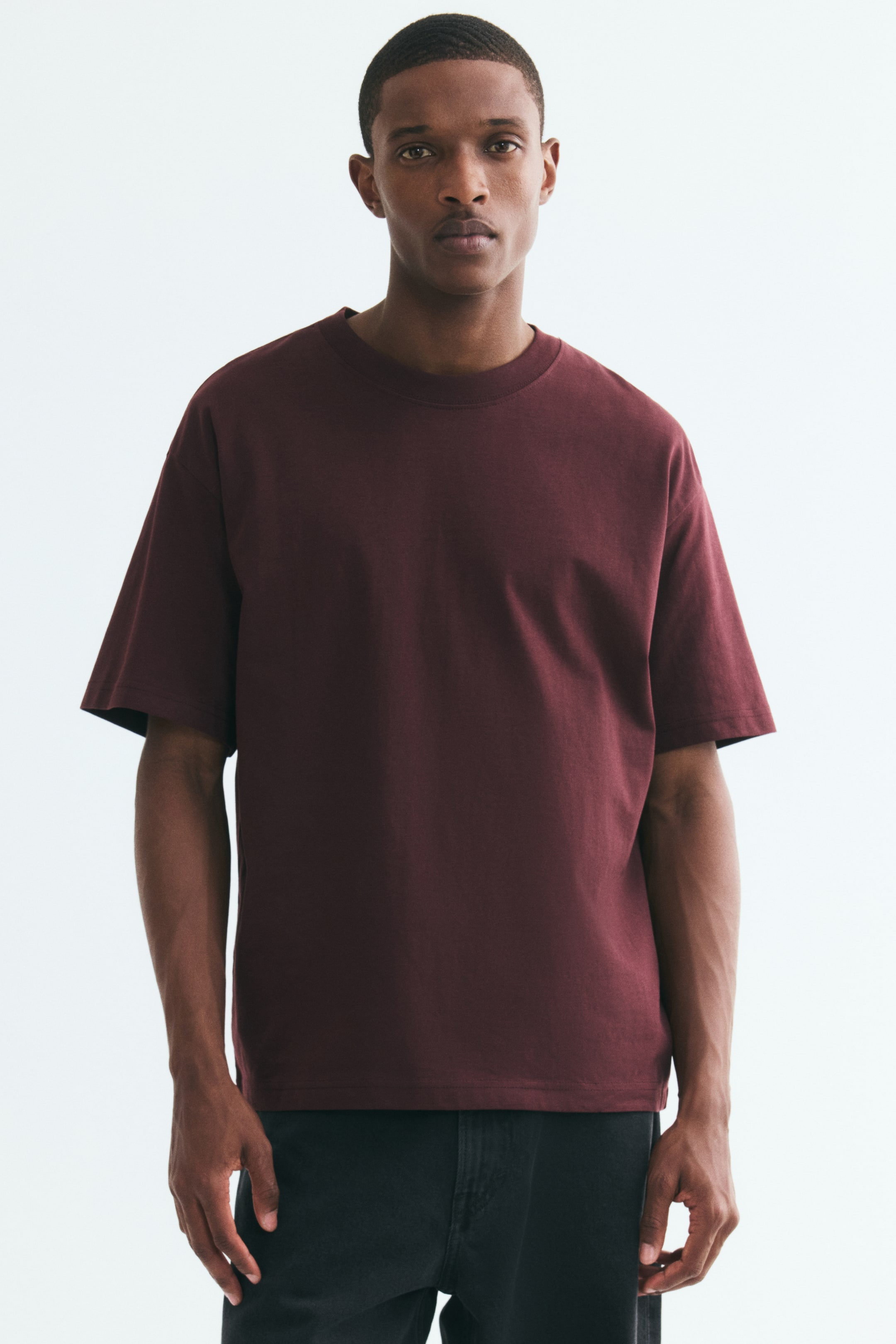 View larger image: The model is wearing a burgundy midweight cotton jersey T-shirt, featuring a relaxed fit, a round rib-trimmed neckline, dropped shoulders, and a straight hem. The T-shirt is styled with dark-coloured trousers.