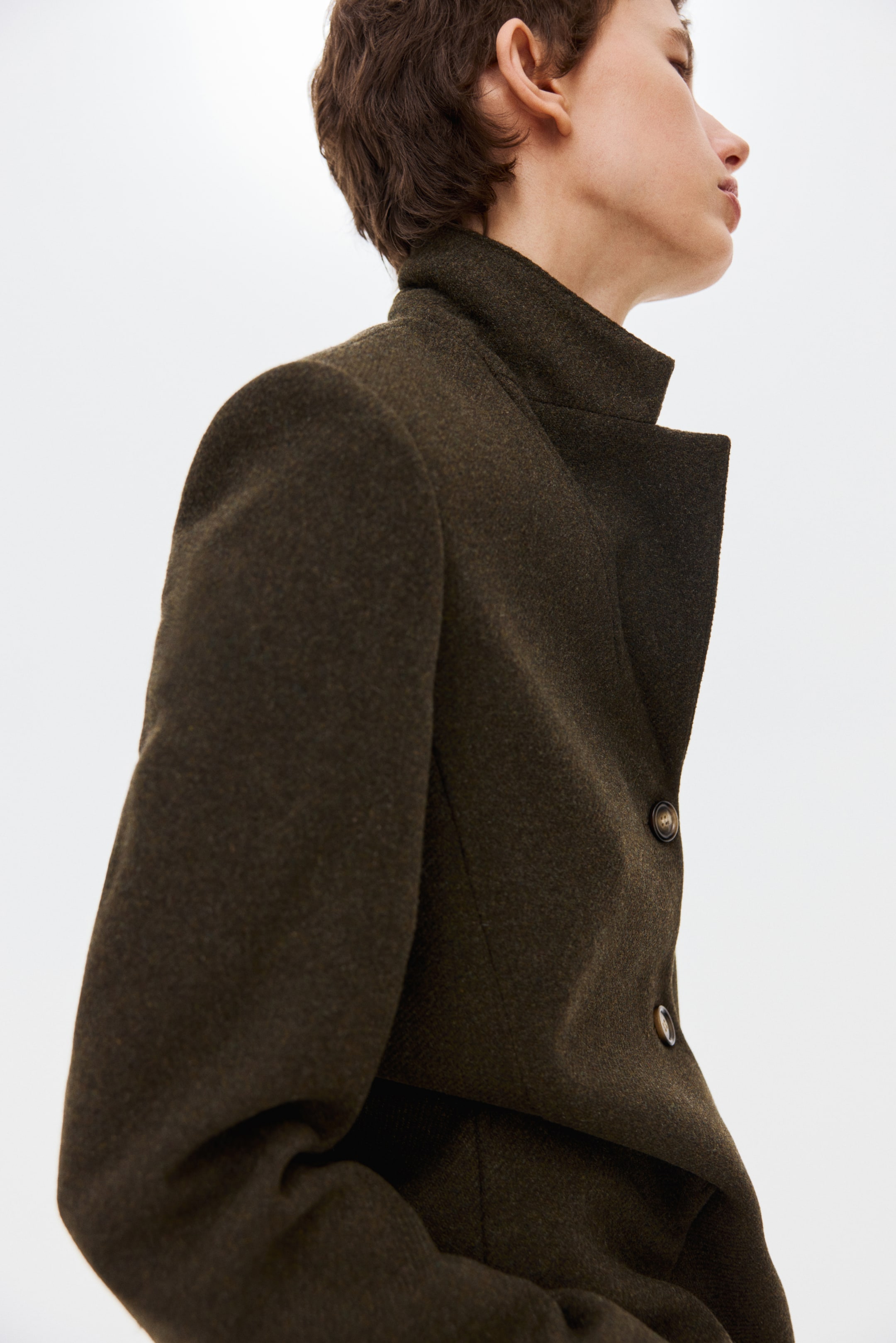 Ampliar la imagen: Una chaqueta texturizada en verde oliva oscuro presenta un cuello alto y dos botones frontales visibles. Se muestra con un corte entallado desde un ángulo lateral-frontal, resaltando su hombro estructurado.