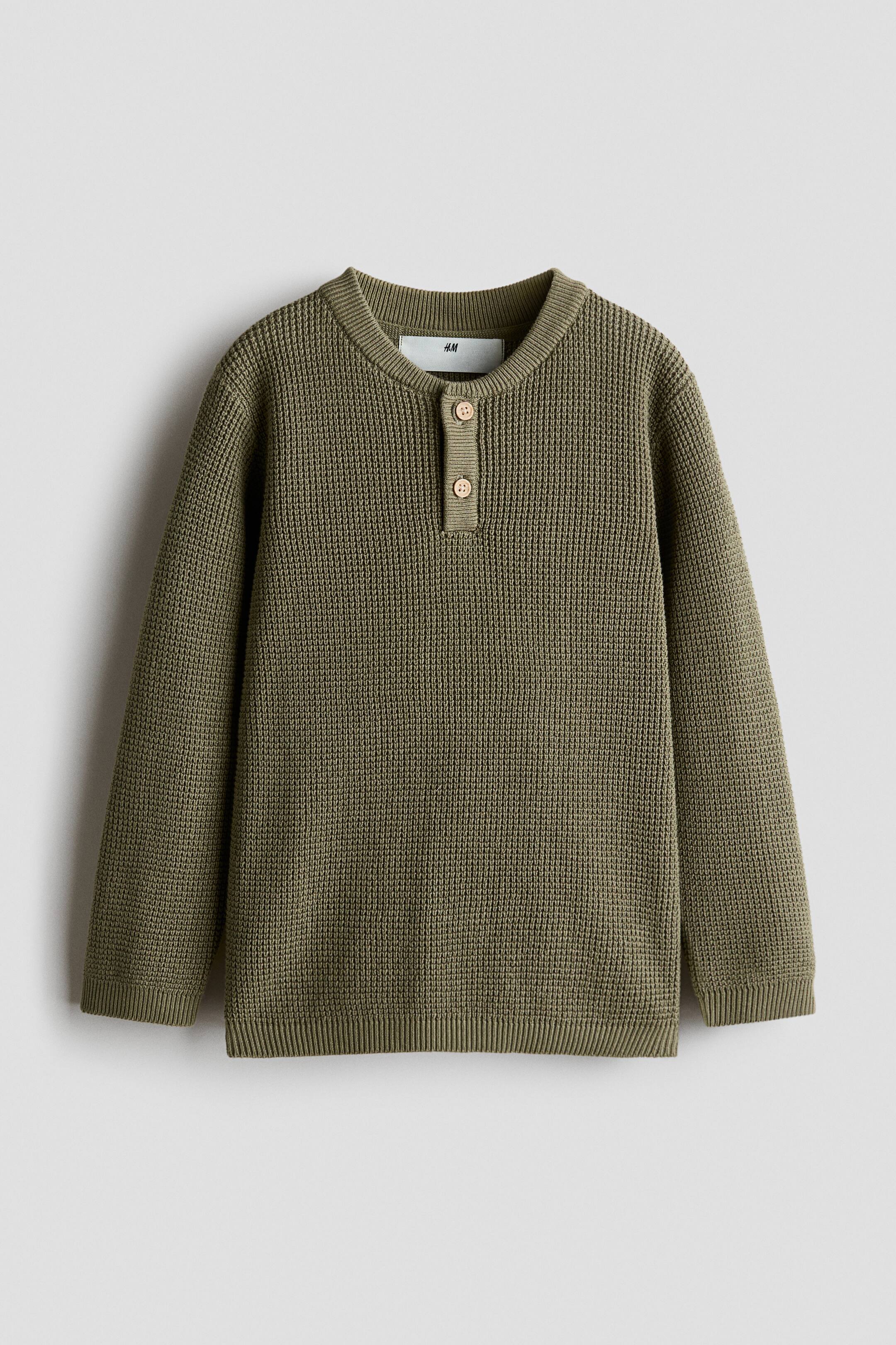 View larger image: An olive green jumper is shown from the front, featuring a waffle knit texture. It is designed with a ribbed Henley collar that includes two light brown wooden buttons, dropped shoulders, and ribbing at the cuffs and hem.