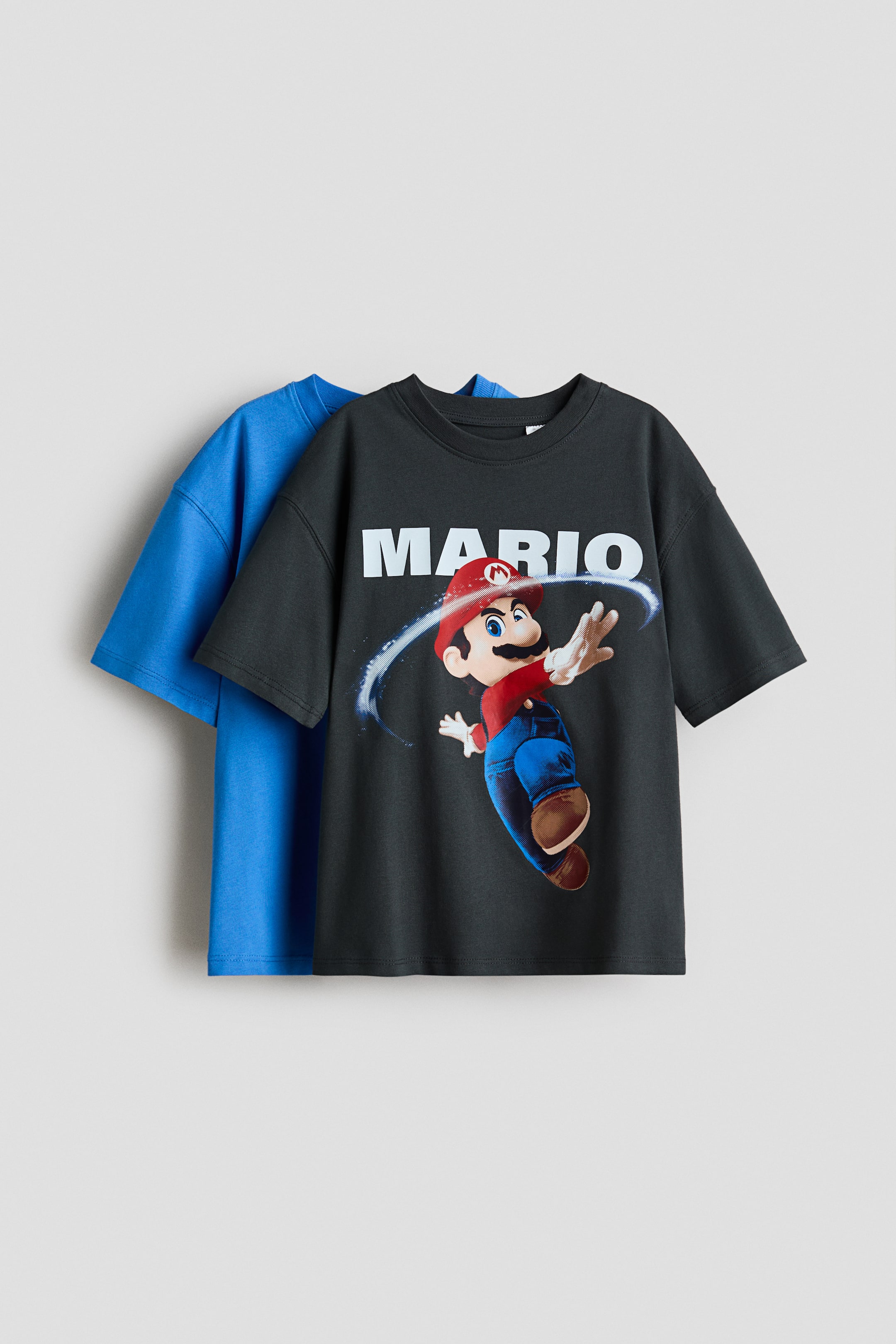 Größeres Bild anzeigen: Ein dunkelgraues, locker sitzendes T-Shirt aus weichem Baumwolljersey präsentiert seine Vorderseite, mit einem 'MARIO'-Print und der Figur Mario, einem rund gerippten Halsausschnitt und überschnittenen Schultern. Ein blaues, locker sitzendes T-Shirt ist dahinter geschichtet und zeigt ebenfalls seine überschnittenen Schultern.