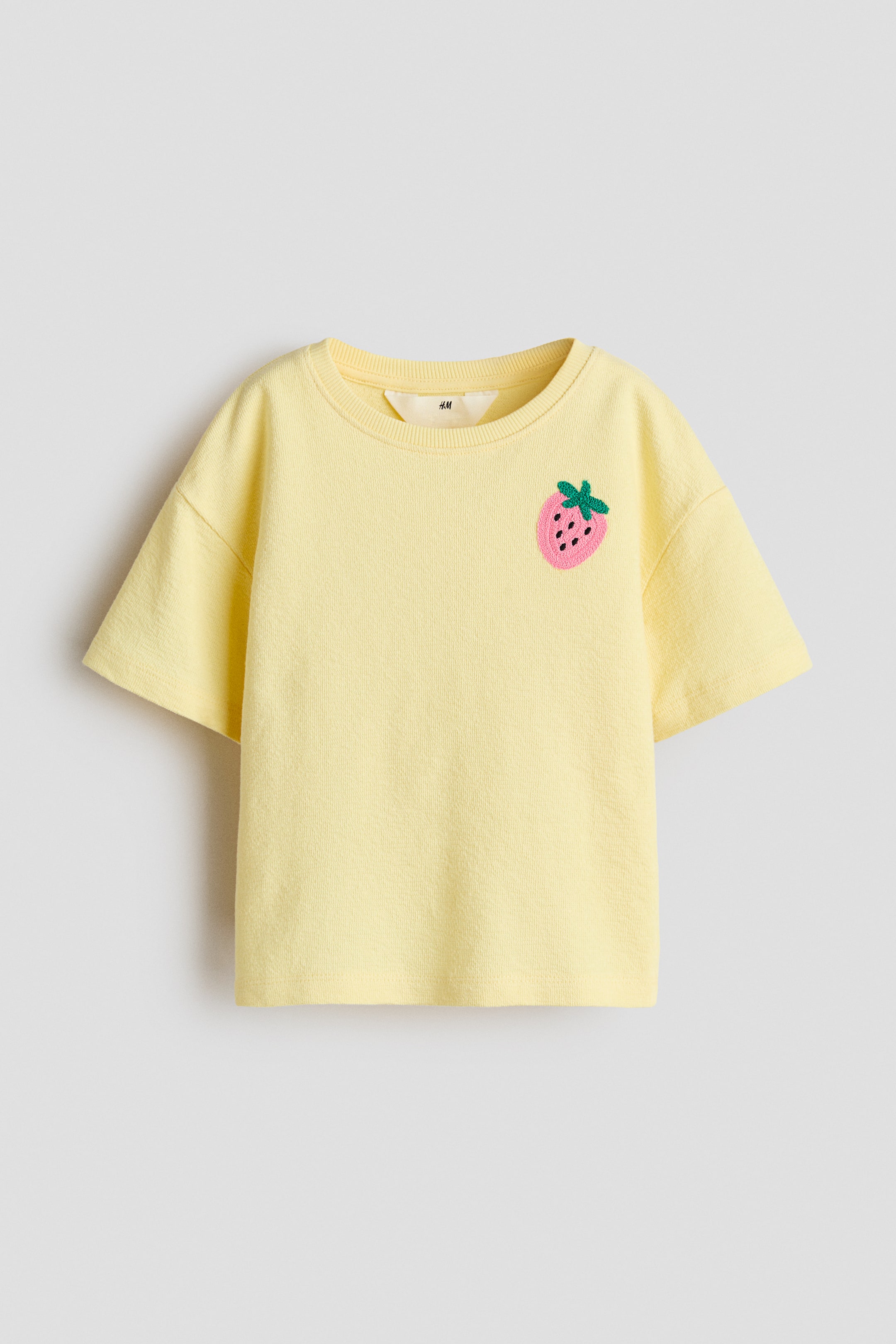Größeres Bild ansehen: Ein gelbes T-Shirt aus strukturiertem Jersey zeigt sich von vorne, mit einem runden, gerippten Ausschnitt, überschnittenen Schultern, kurzen Ärmeln und einer pinkfarbenen Erdbeer-Applikation mit schwarzen Samen und grünem Stiel auf der linken Brust.