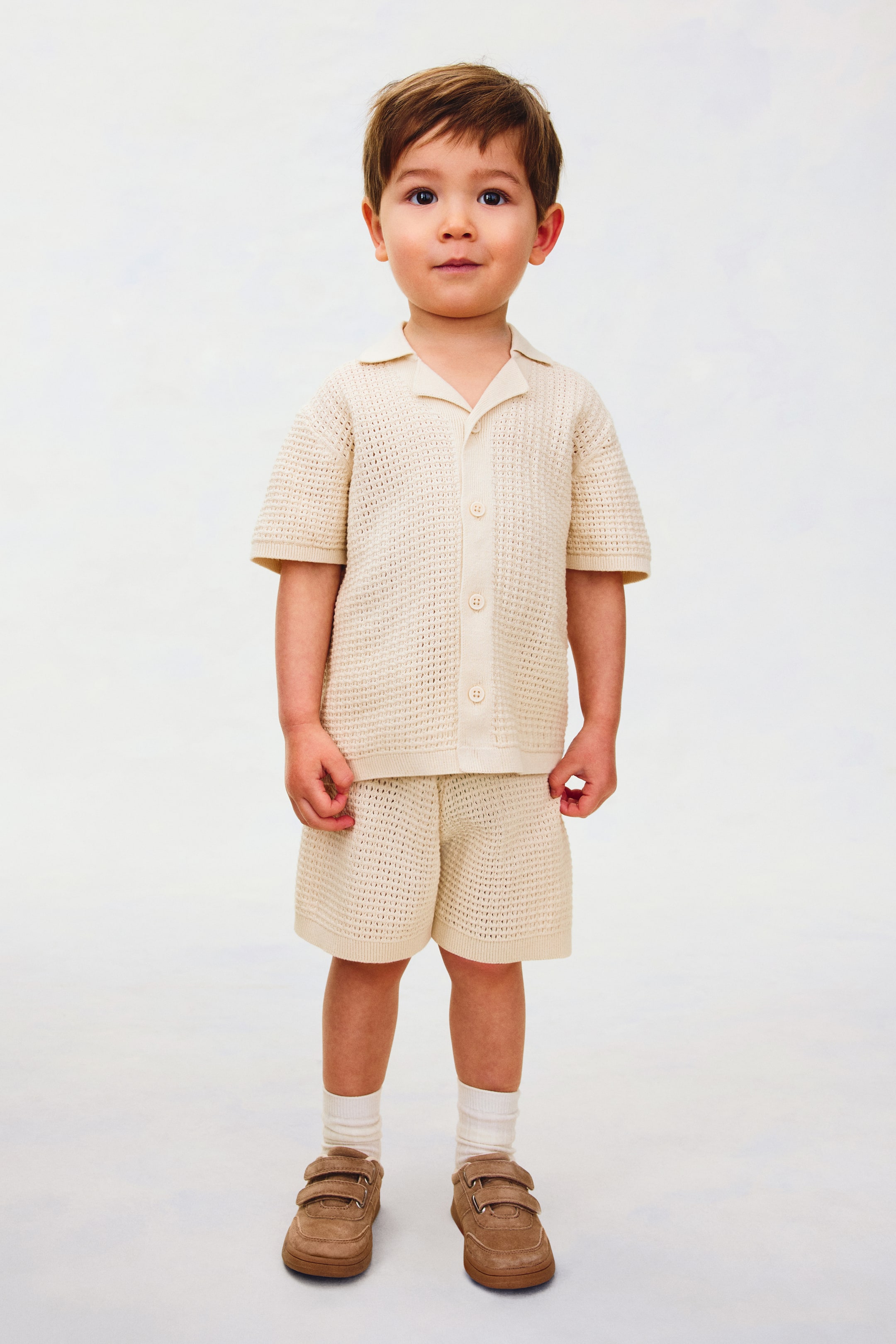 Größeres Bild ansehen: Ein hellbeiges, offen gestricktes Zweiteiler-Set wird präsentiert, bestehend aus einem kurzärmeligen Hemd mit einem offenen Kragen und fünf Knöpfen vorne, sowie passenden Shorts.