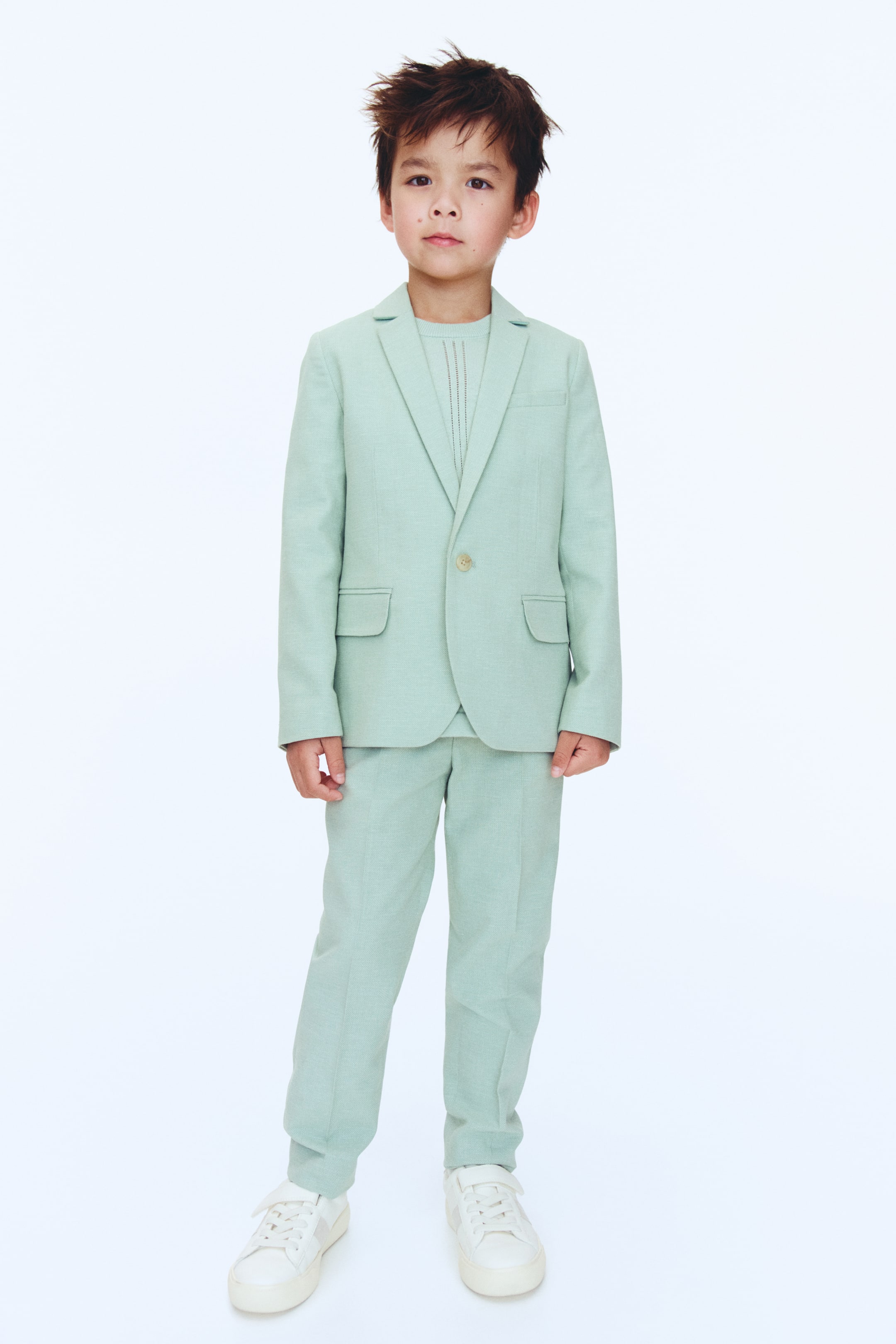 View larger image: A light green child's suit, crafted from a textured weave fabric, is on display. The single-breasted blazer boasts notch lapels, a single light-coloured button fastening, and two flap pockets. Complementing the ensemble are matching straight-leg trousers, while a light green jumper with a vertical textured knit is worn underneath the blazer.