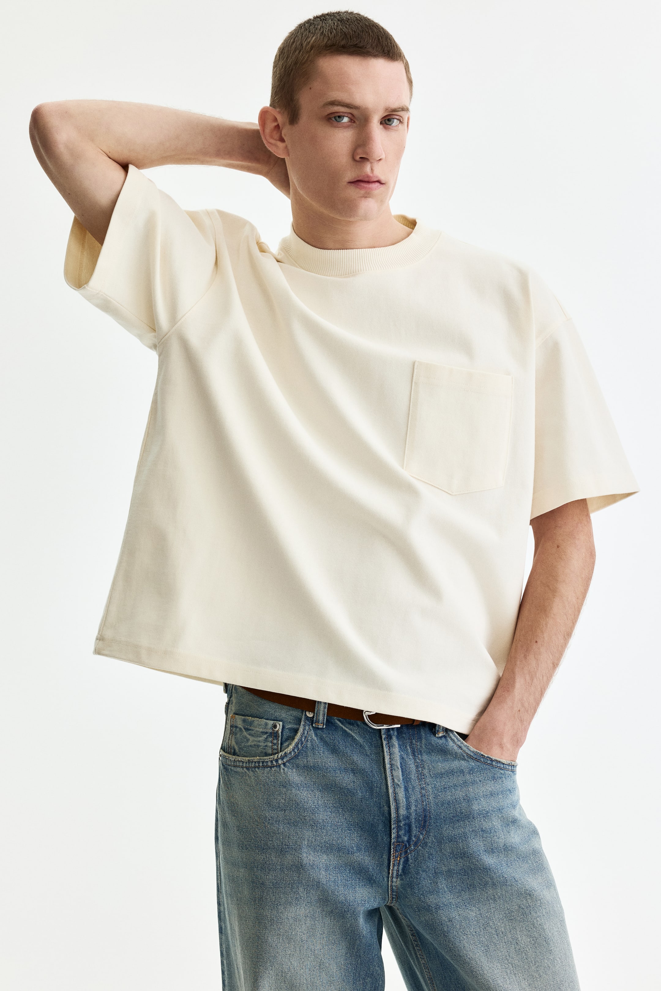 View larger image: The cream t-shirt features a loose, boxy fit with dropped shoulders and short, wide sleeves. It has a ribbed crewneck and a single patch pocket on the left chest.
