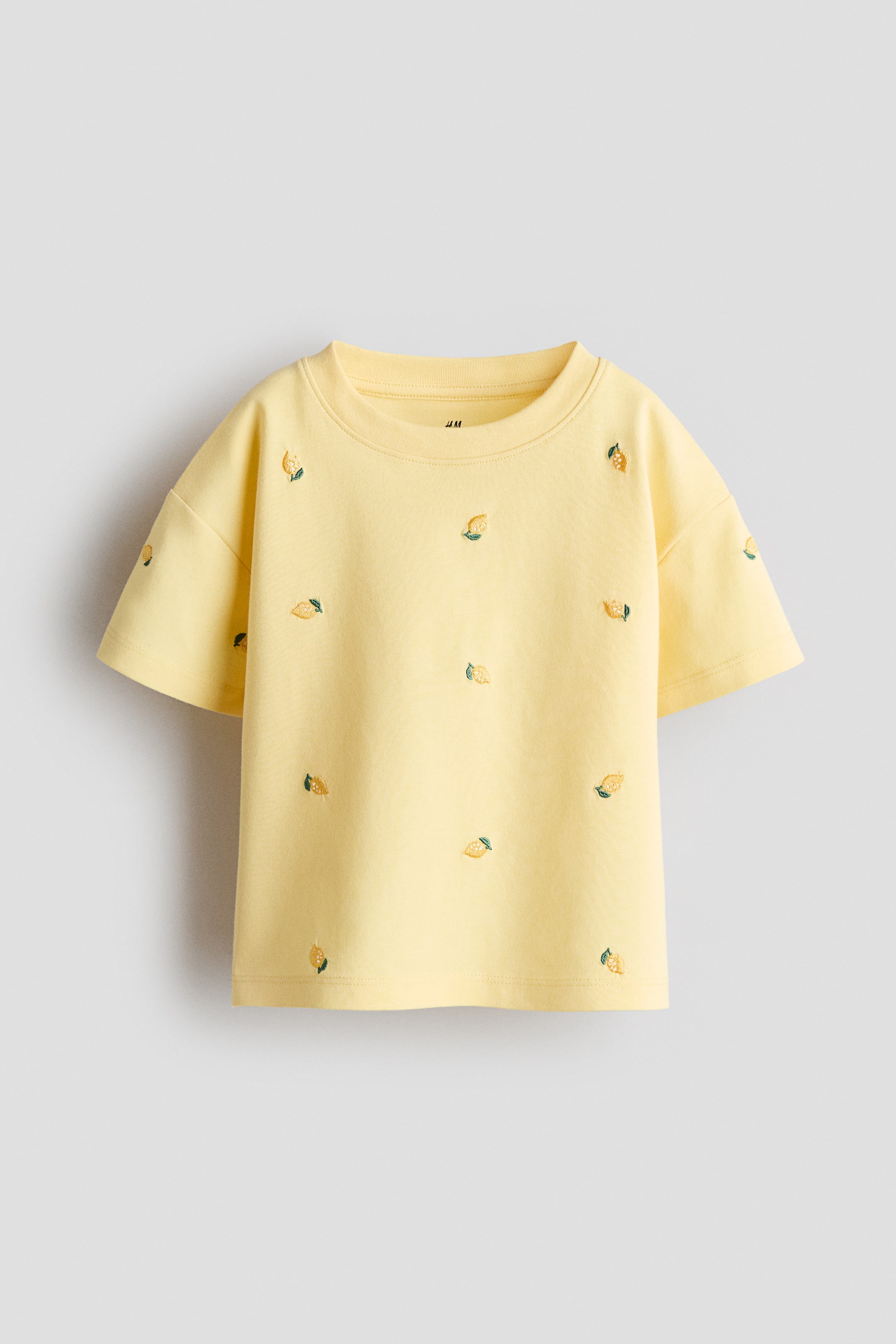 Grotere afbeelding bekijken: Een T-shirt met een relaxte pasvorm in lichtgeel wordt frontaal gepresenteerd, met een geribbelde afwerking rond de halslijn en laag ingezette schouders. Het is gemaakt van zachte katoenjersey en is versierd met kleine geborduurde citroenmotieven die over het oppervlak verspreid zijn.
