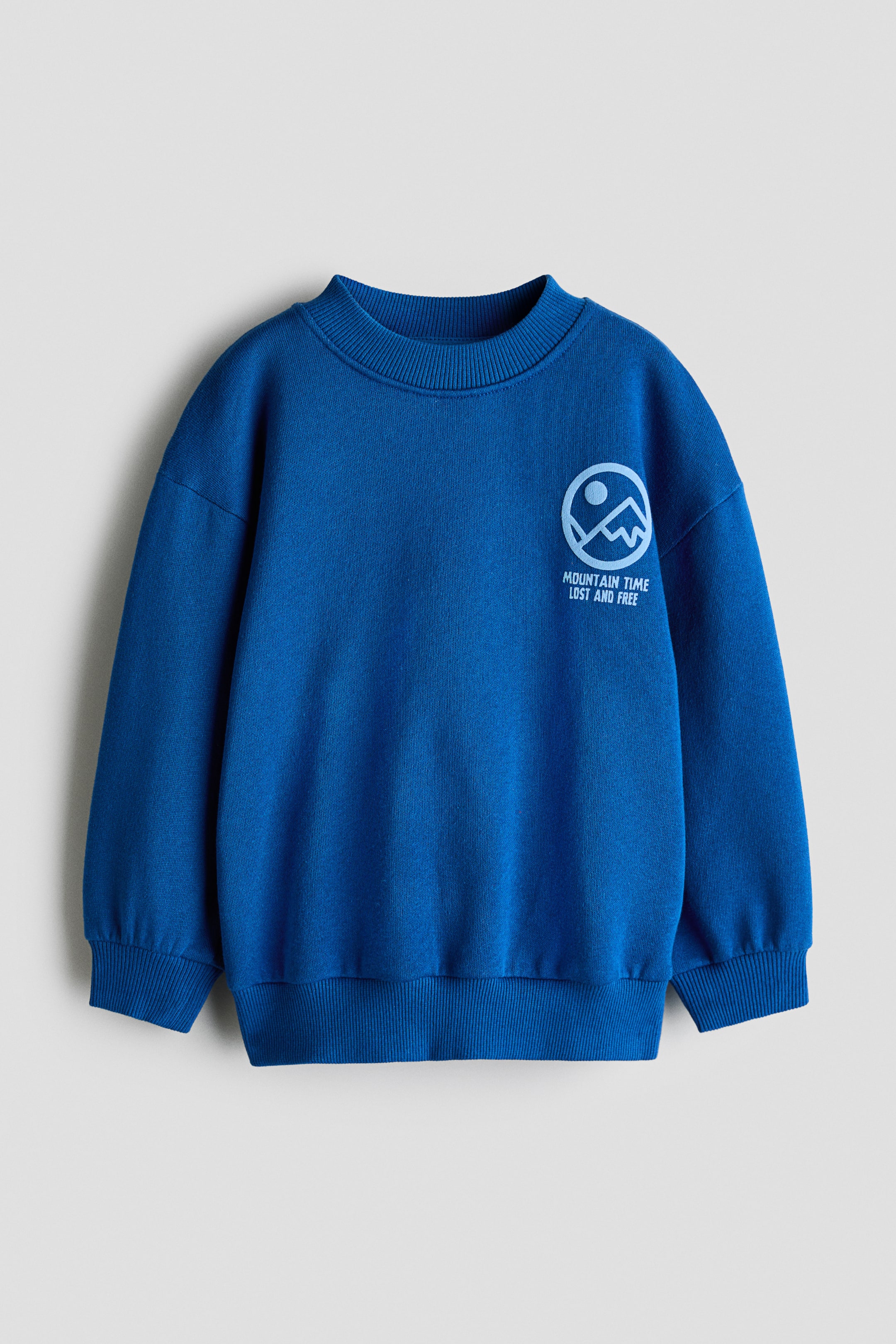 Grotere afbeelding bekijken: Een blauwe sweatshirt is van voren te zien en toont een ronde halslijn met ribboord, verlaagde schouders en lange mouwen met geribde boorden. Op de linkerborst is een lichtblauwe graphic print zichtbaar met een cirkelvormig bergontwerp en de tekst "MOUNTAIN TIME LOST AND FREE" boven de geribde zoom.