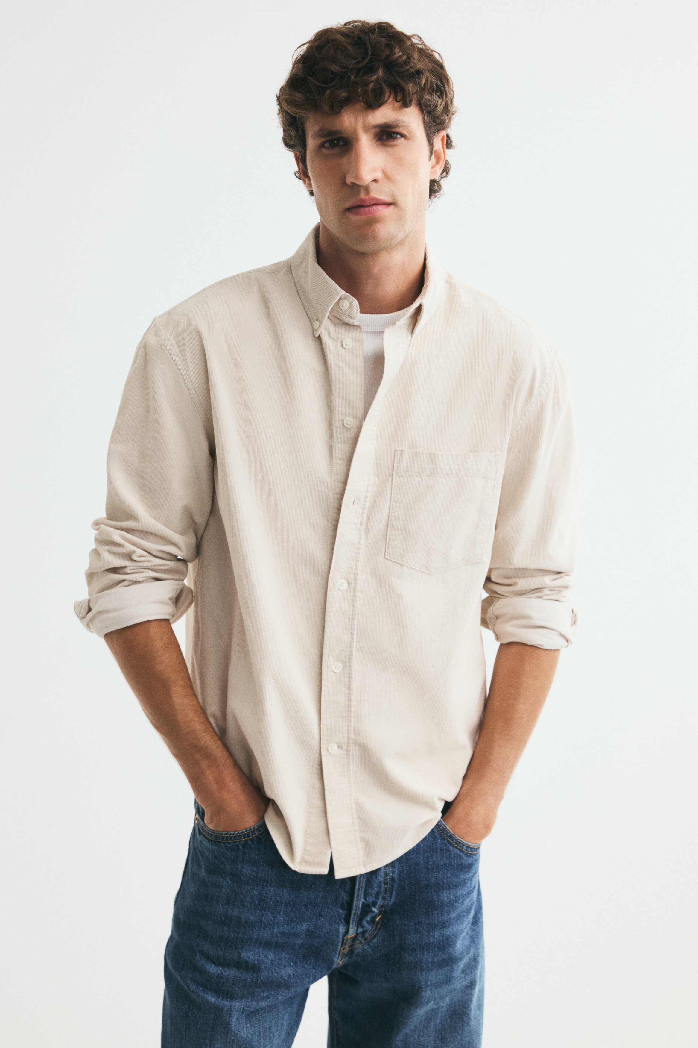 Una camisa de pana en color beige claro se presenta en vista frontal, con un cuello abotonado, una tapeta de botones central y un único bolsillo en el pecho abierto. Está diseñada con mangas largas, puños ajustables con botones, tapetas en las mangas y un dobladillo redondeado.