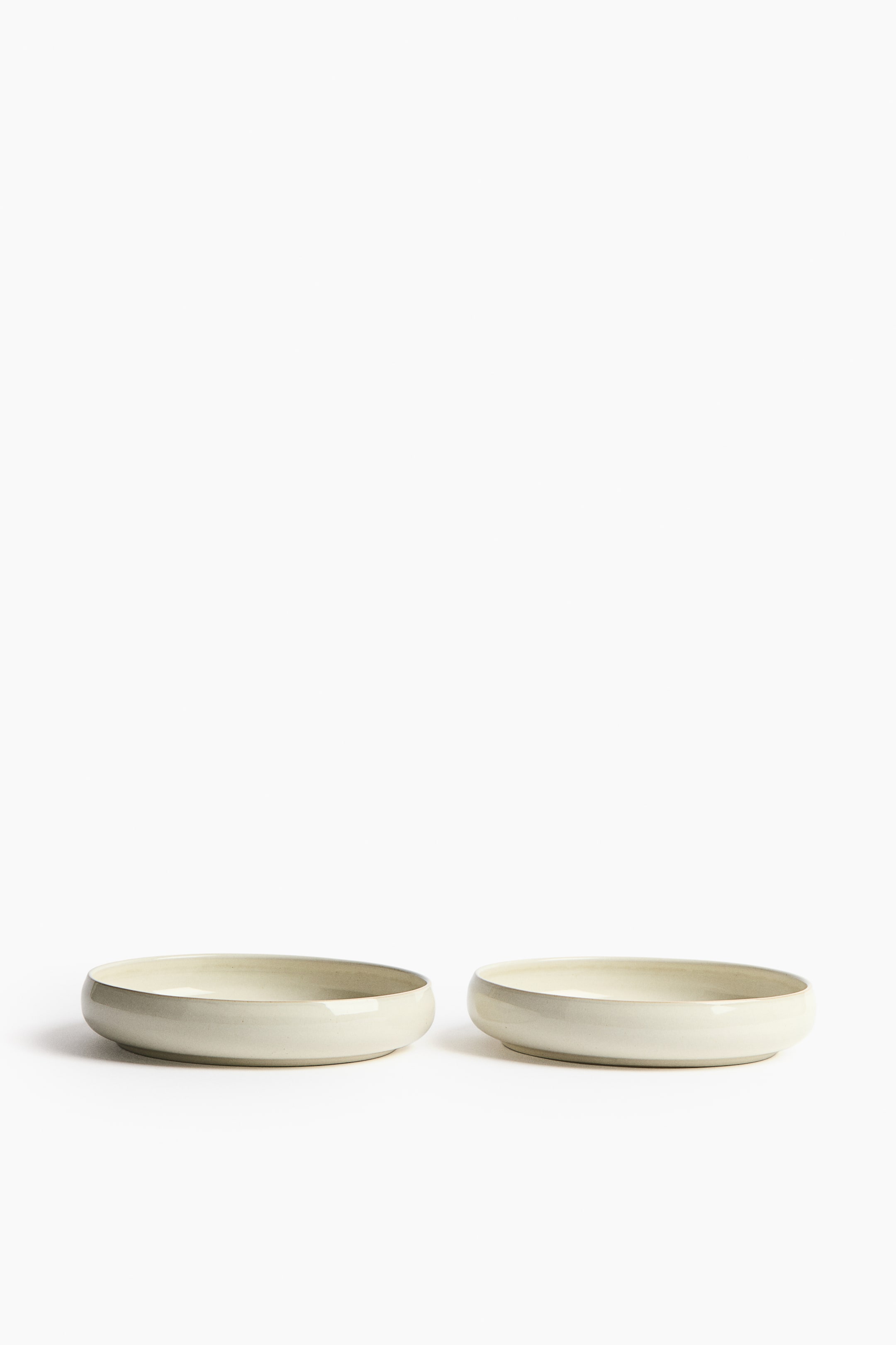 View larger image: Two shallow, creamy off-white ceramic bowls are presented from a slightly angled front view. They feature wide, rounded forms and smooth, glossy surfaces.