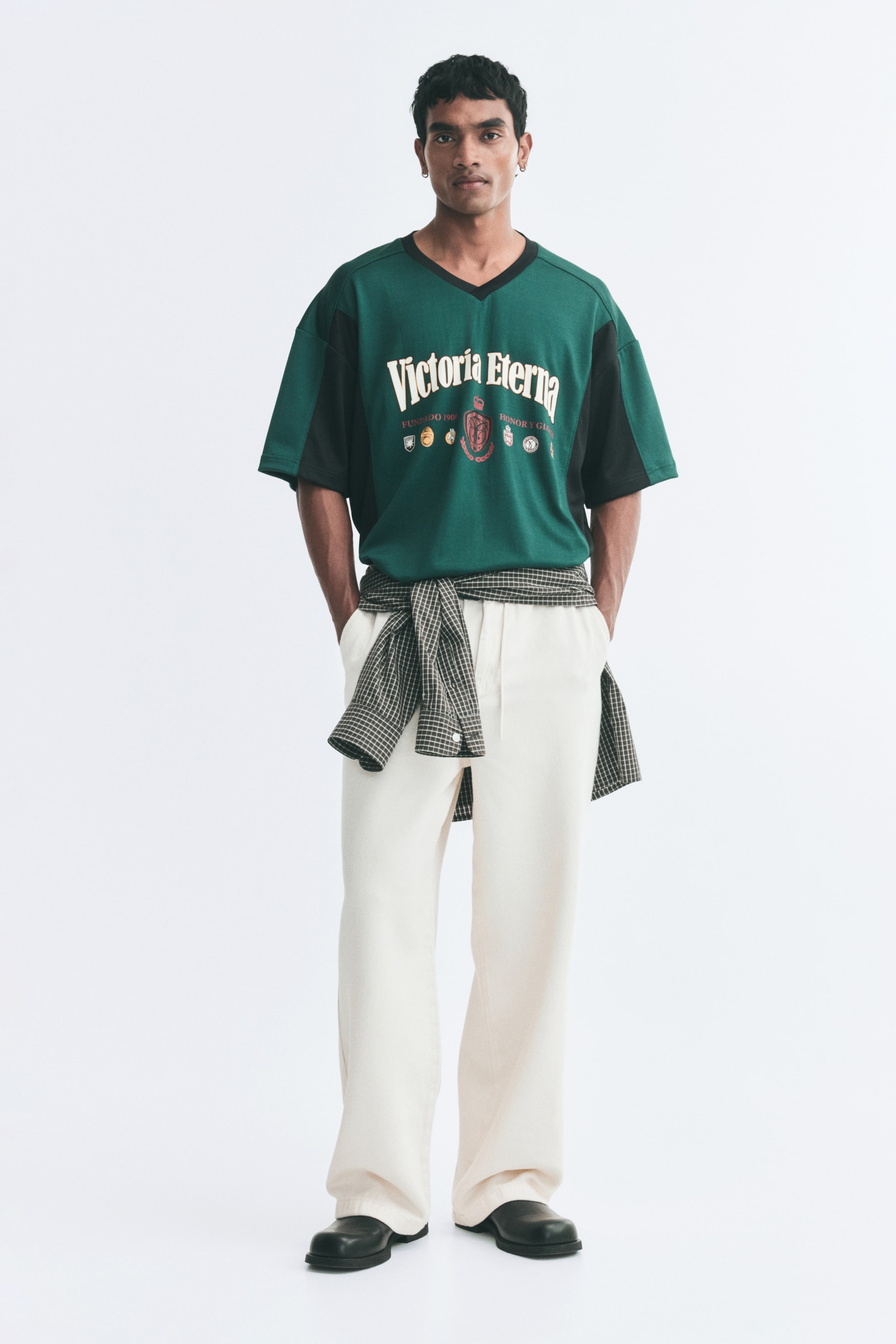 View larger image: A relaxed-fit jersey T-shirt in green and black, featuring a rib-trimmed V-neck and printed graphic designs on the front, is worn by the model. The T-shirt is styled with cream-coloured wide-leg trousers and a plaid shirt tied around the waist.