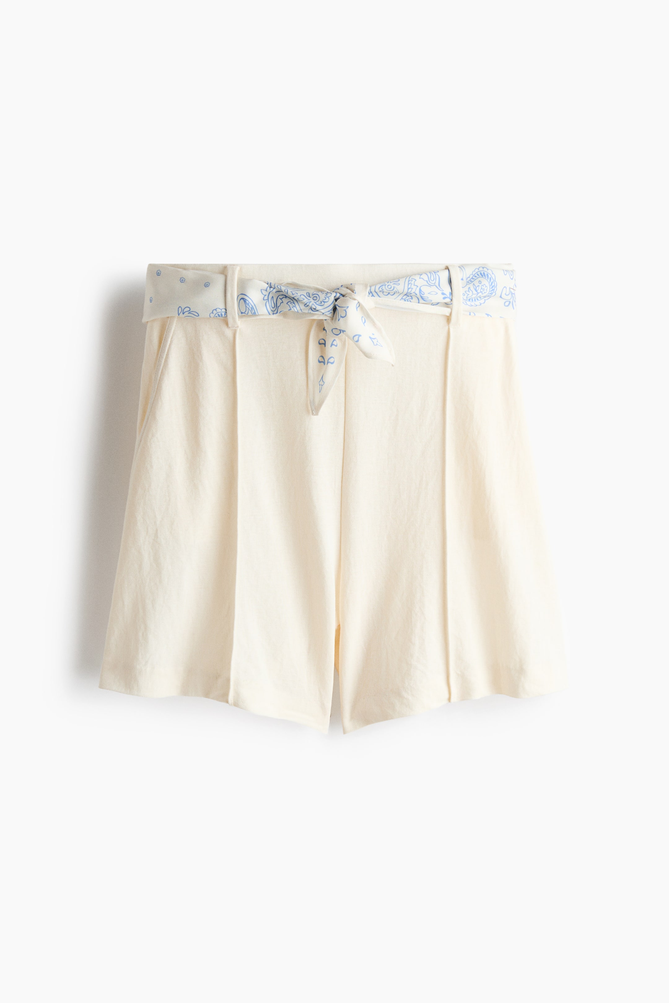 Größeres Bild ansehen: "Off-White Shorts präsentieren eine Vorderansicht mit hoher Taille, Gürtelschlaufen und sichtbaren Seitentaschen. Ein weißer Stoffgürtel mit blauen Paisley-Mustern ist durch die Schlaufen gezogen und vorne zu einem Knoten gebunden, mit vertikalen Falten, die sich über die Vorderseite der Shorts erstrecken."
