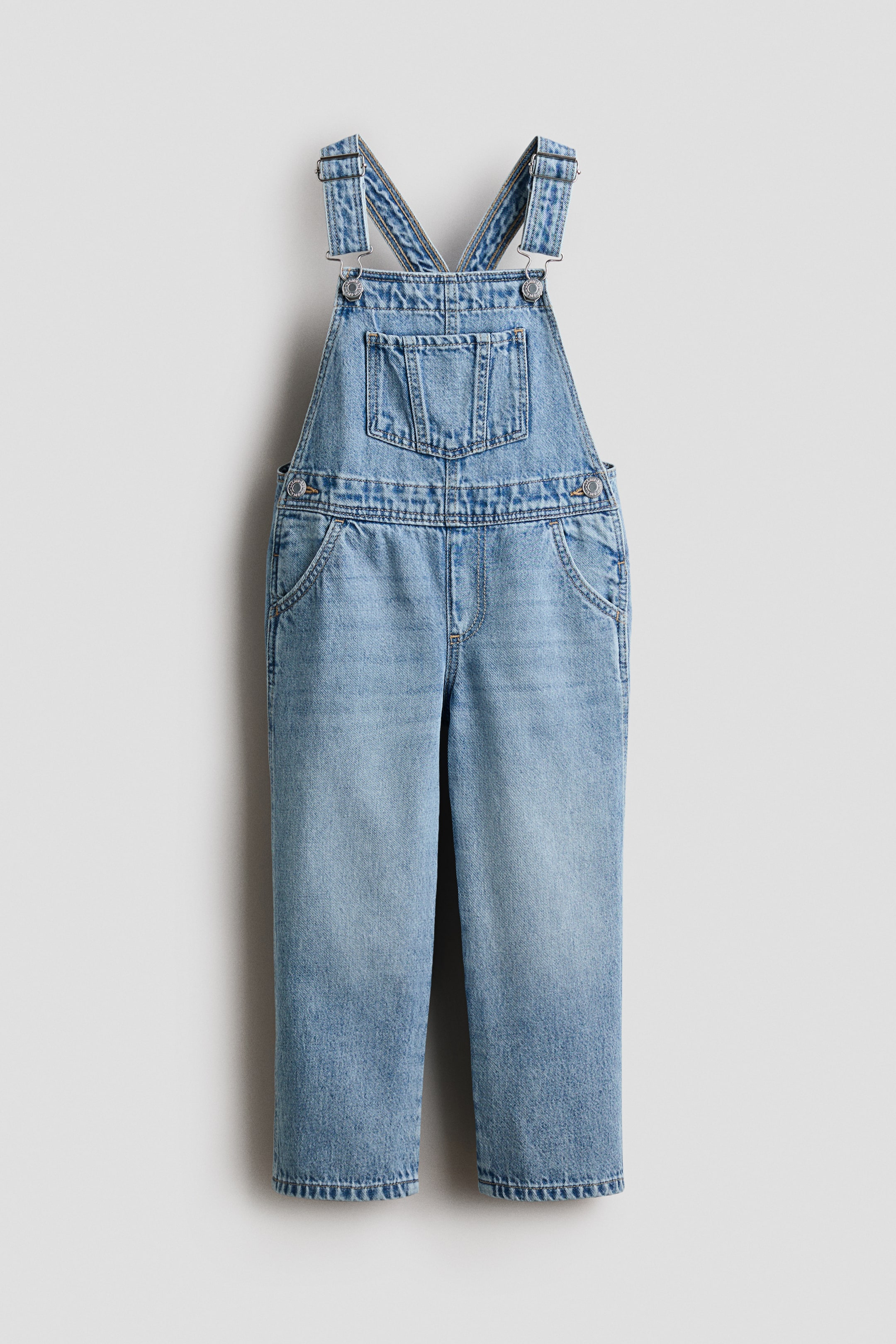 Se større billede: Overall i lyseblå vasket bomuldsdenim præsenteres forfra, hvor justerbare stropper krydser øverst, trykknapper i siderne og en falsk gylp er synlige. En åben brystlomme og to forlommer er synlige.
