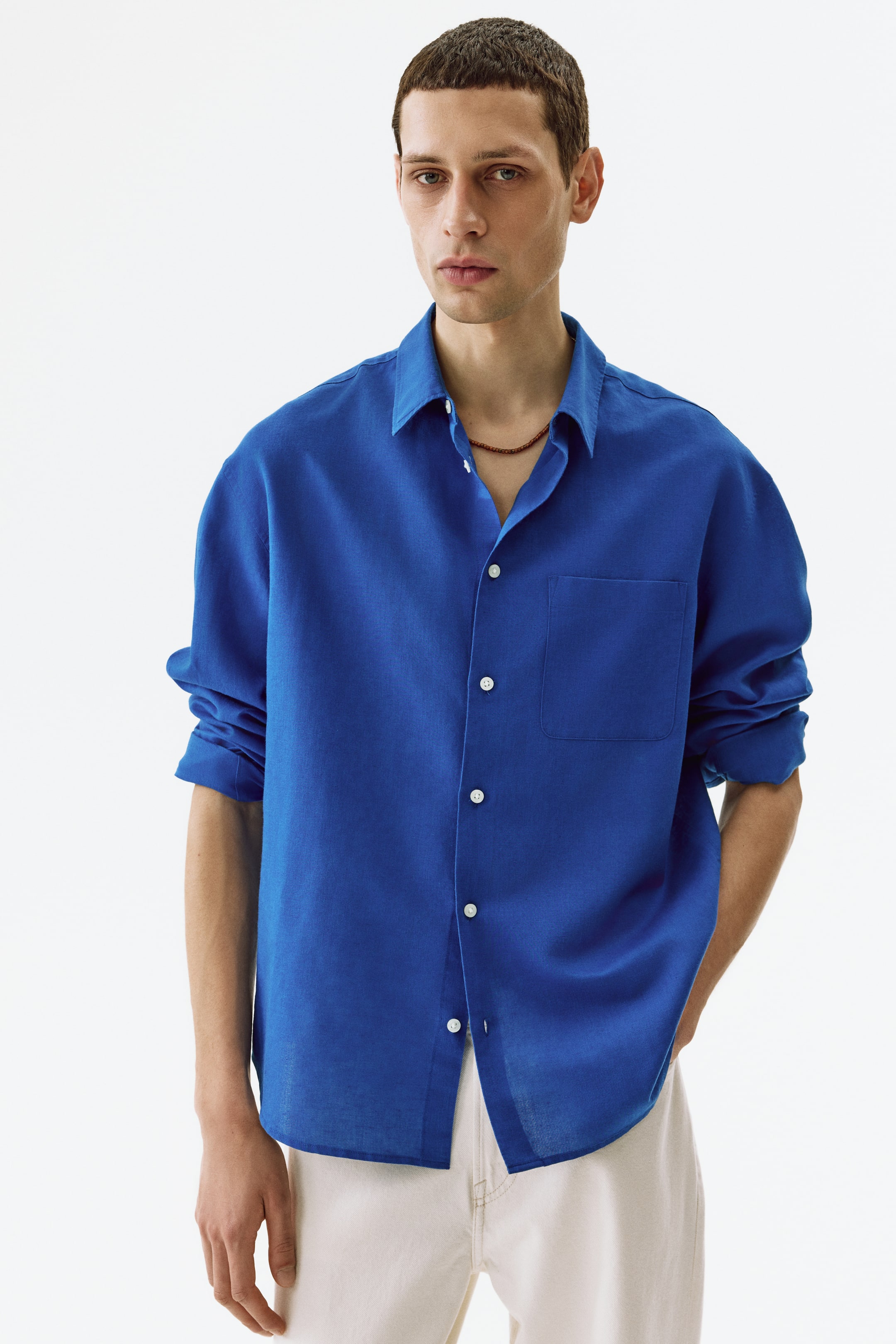 View larger image: A royal blue button-up shirt is displayed with a relaxed fit and textured fabric, shown facing forward. It features a spread collar, a left chest patch pocket, and white buttons running down the front. The long sleeves are rolled up, and the top buttons are left undone, revealing a dark beaded necklace.