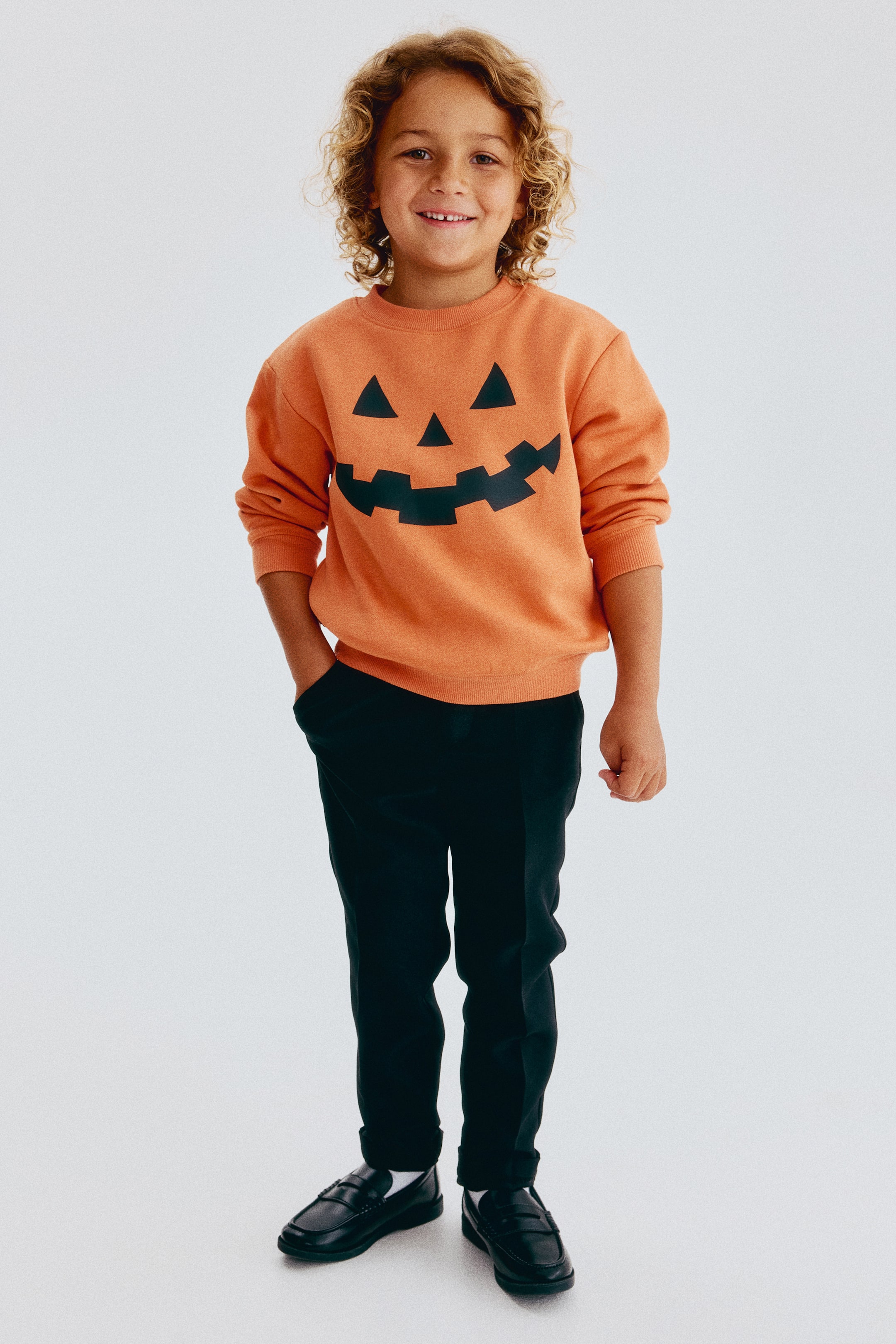 View larger image: An orange sweatshirt made from sweat fabric features a printed black jack-o'-lantern design on the front and is shown on a child. The sweatshirt has a crew neckline with ribbed trim, dropped shoulders, long sleeves with ribbed cuffs, and a ribbed hem.