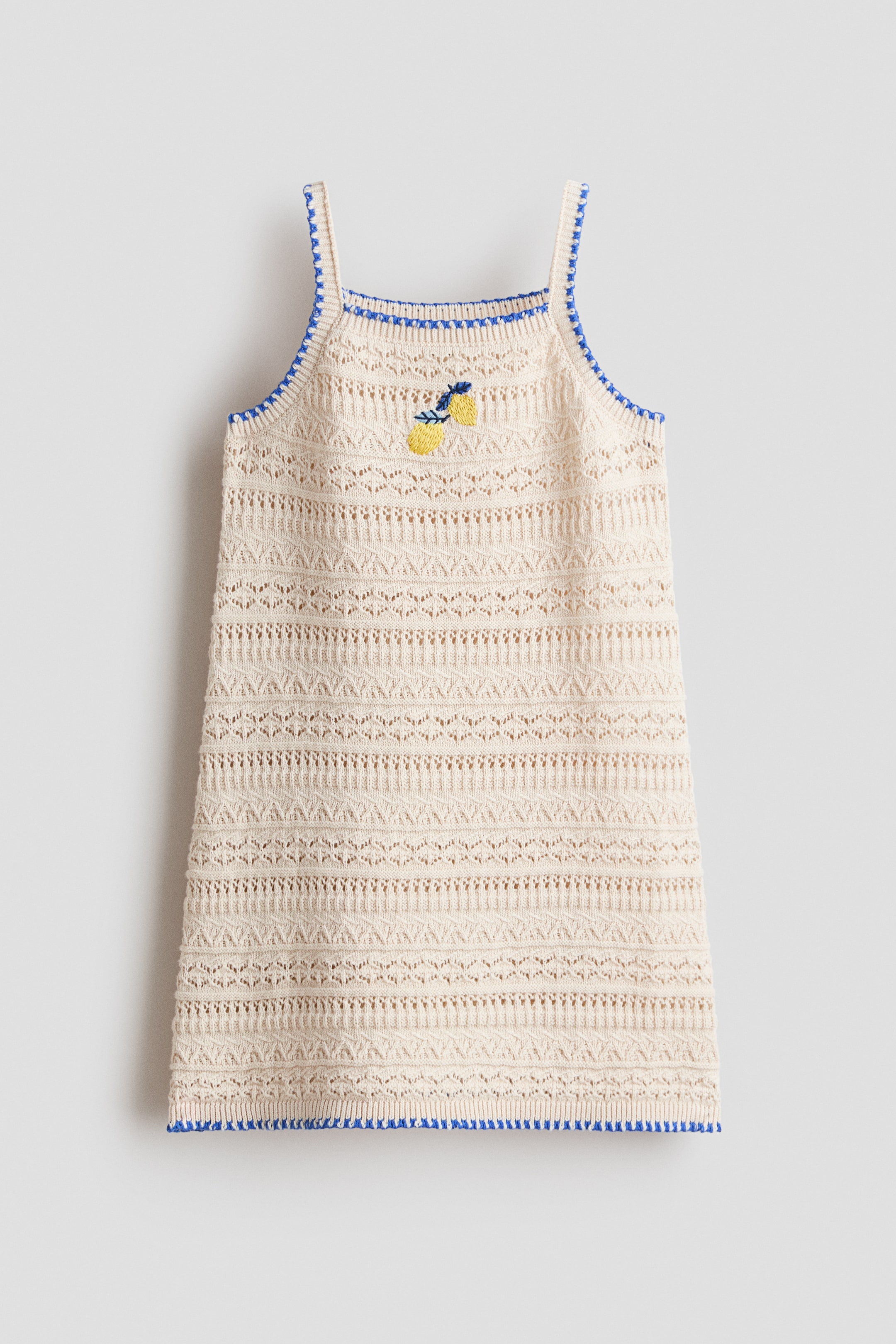 View larger image: The cream-coloured sleeveless knit dress is displayed front-facing, featuring blue trim along the neckline, straps, and hem. It showcases horizontal bands of openwork patterns throughout its body, with two yellow lemon embroideries adorning the chest.