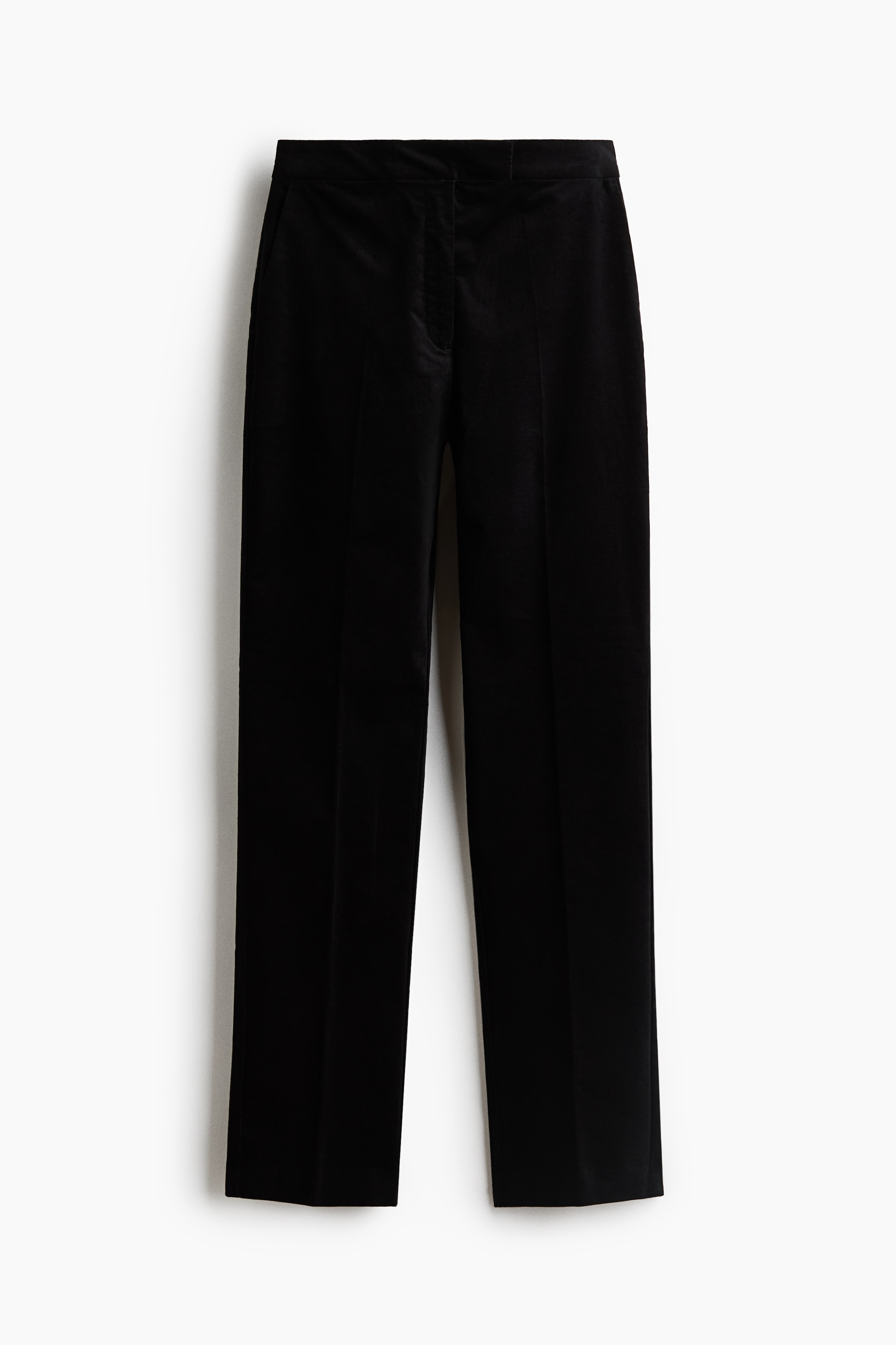 Black fitted velvet pants featuring slim legs with front creases are being worn by the model, who is standing with hands resting in the diagonal side pockets. The pants are styled with a black long-sleeve sweater layered over a white top and paired with black heeled sandals.