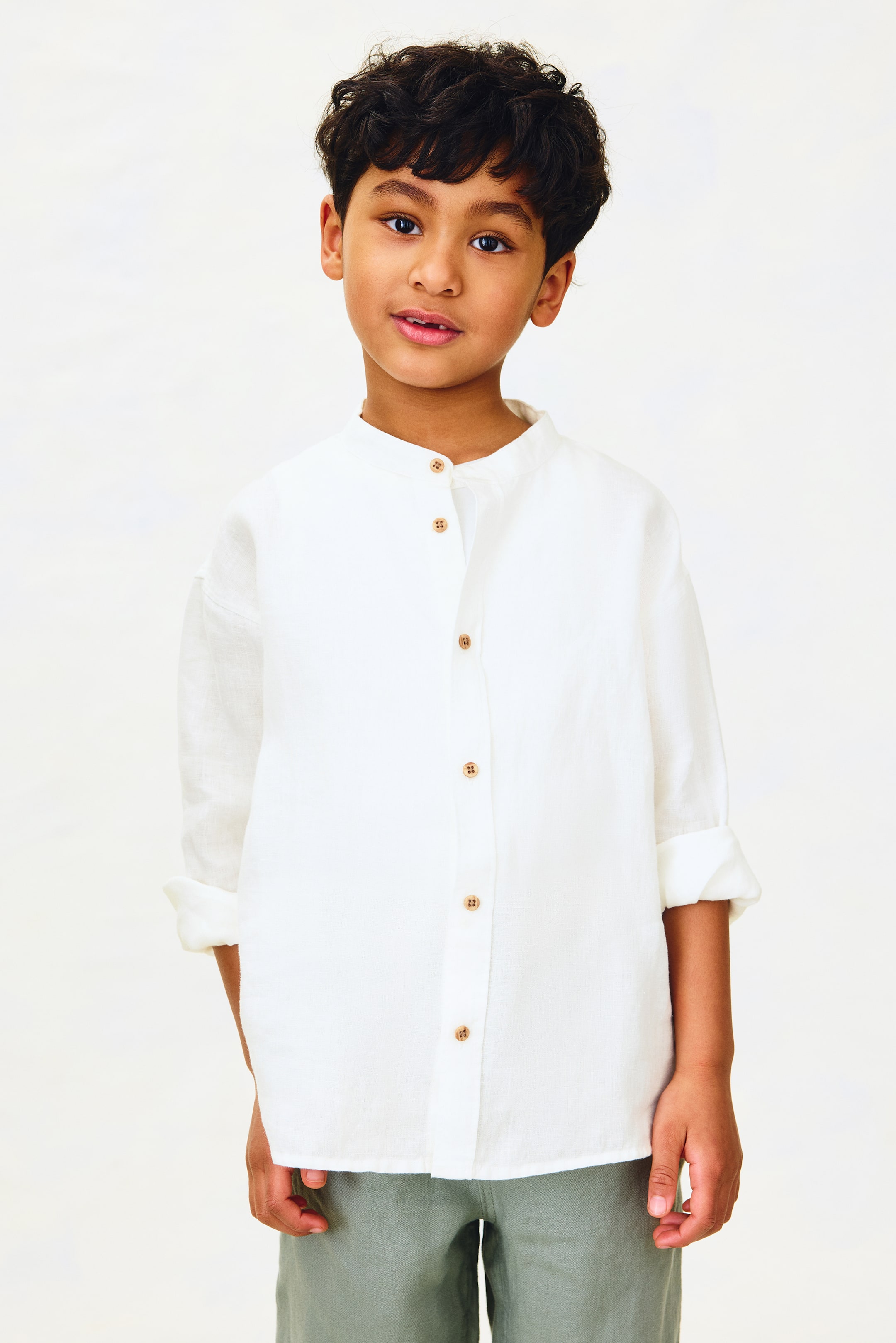 Visualizza immagine più grande: Una camicia bianca presenta un colletto alla coreana e una mezza abbottonatura con cinque bottoni marrone chiaro. Le maniche lunghe sono arrotolate fino agli avambracci e il tessuto strutturato appare leggermente ampio.