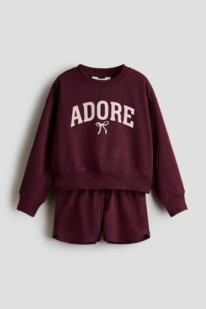 A burgundy sweatshirt and matching sweatshorts set is shown from the front, featuring a long-sleeved top with dropped shoulders, a ribbed crew neckline, ribbed cuffs, and the word "ADORE" printed in light pink above a small bow graphic. The coordinating shorts feature an elasticized drawstring waistband and curved side hems. - 1