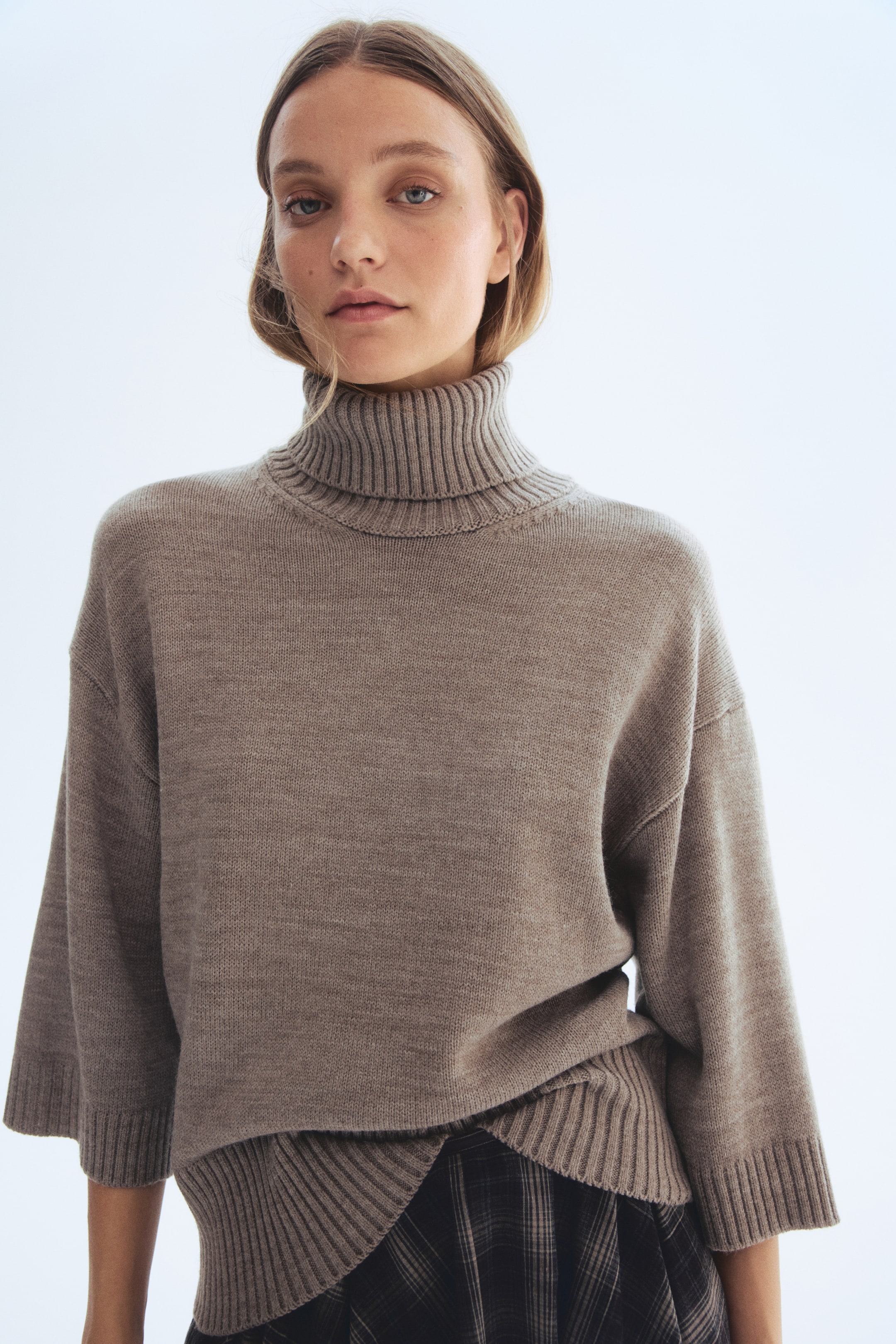 Größeres Bild anzeigen: Ein taupefarbener Strickpullover wird getragen, der mit einem gerippten Rollkragen, weiten Dreiviertelärmeln mit gerippten Bündchen und einem gerippten Saum, der über einem karierten Rock liegt, ausgestattet ist.