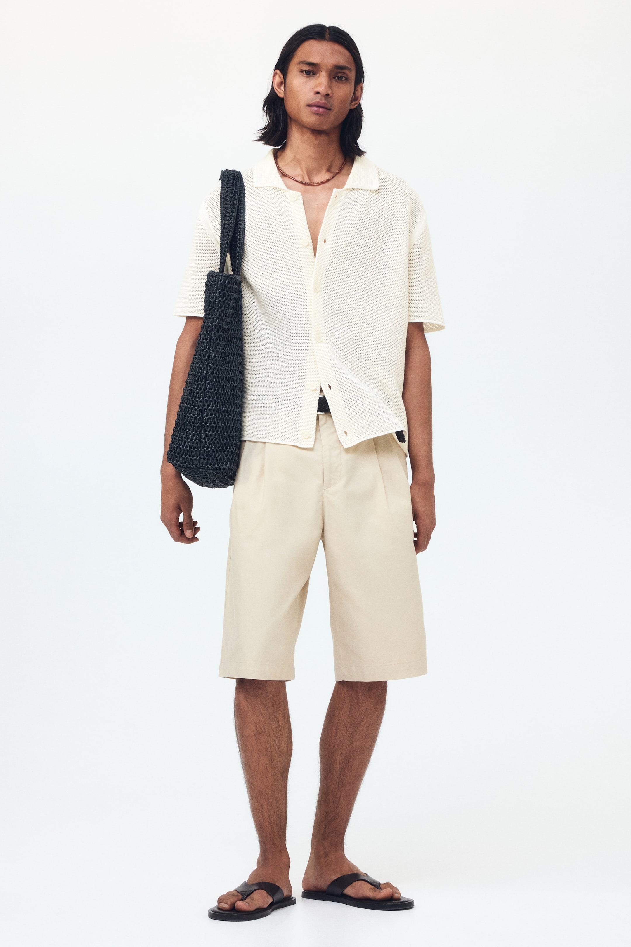 Ver imagen más grande: El conjunto presenta un polo de punto de manga corta en color crema claro, con cuello abierto y cierre de botones en la parte delantera, mostrado de frente y combinado con unos pantalones cortos plisados en beige. Unas sandalias negras de dedo completan el look, mientras que un bolso tote negro tejido se lleva al hombro.