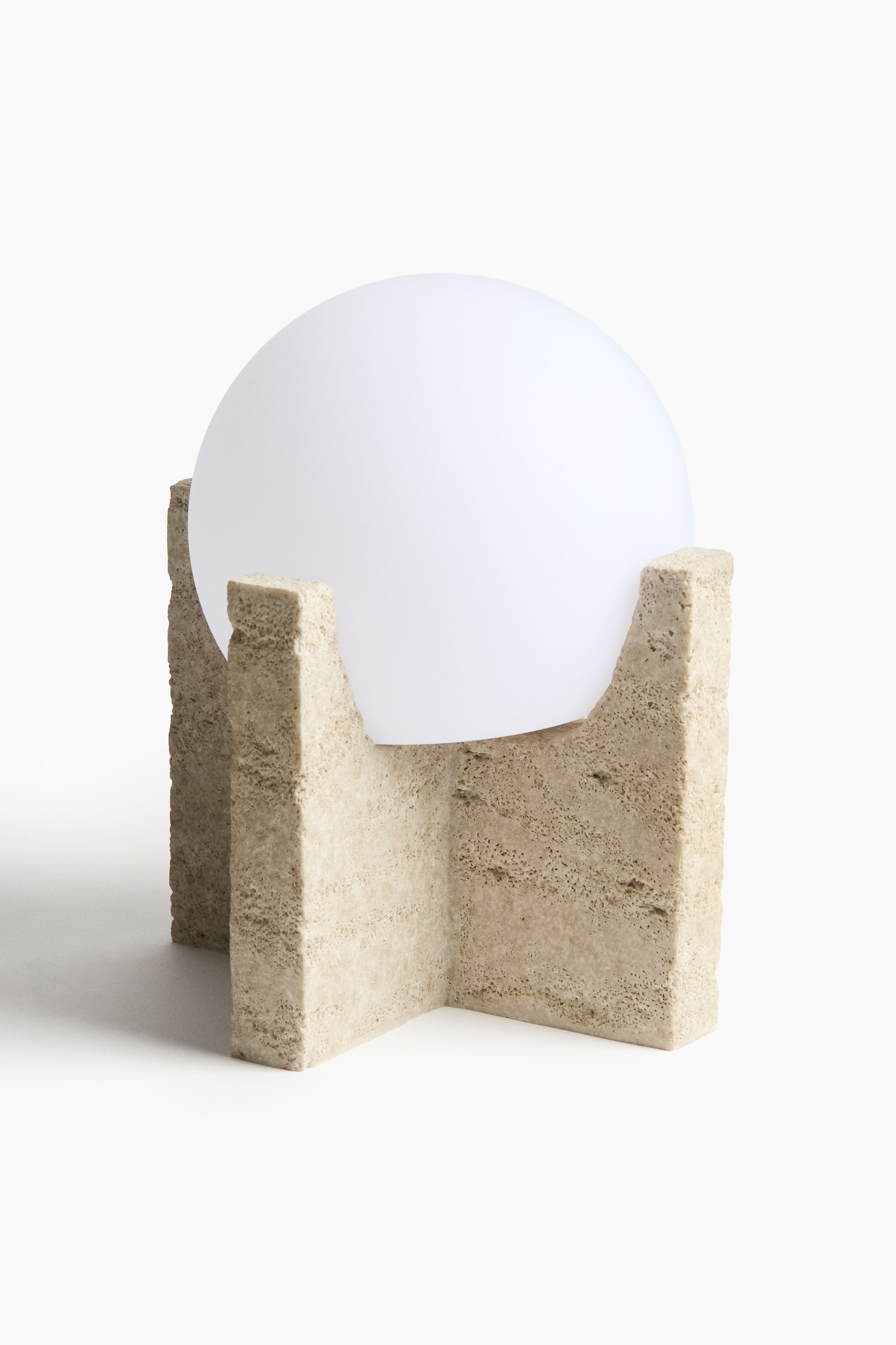 Grotere afbeelding bekijken: Een tafellamp met een bolvormige witte globe die rust in een lichtbeige voet met een ruw oppervlak. De voet vormt een kruisvormige structuur met vier verticale delen die de globe omsluiten.