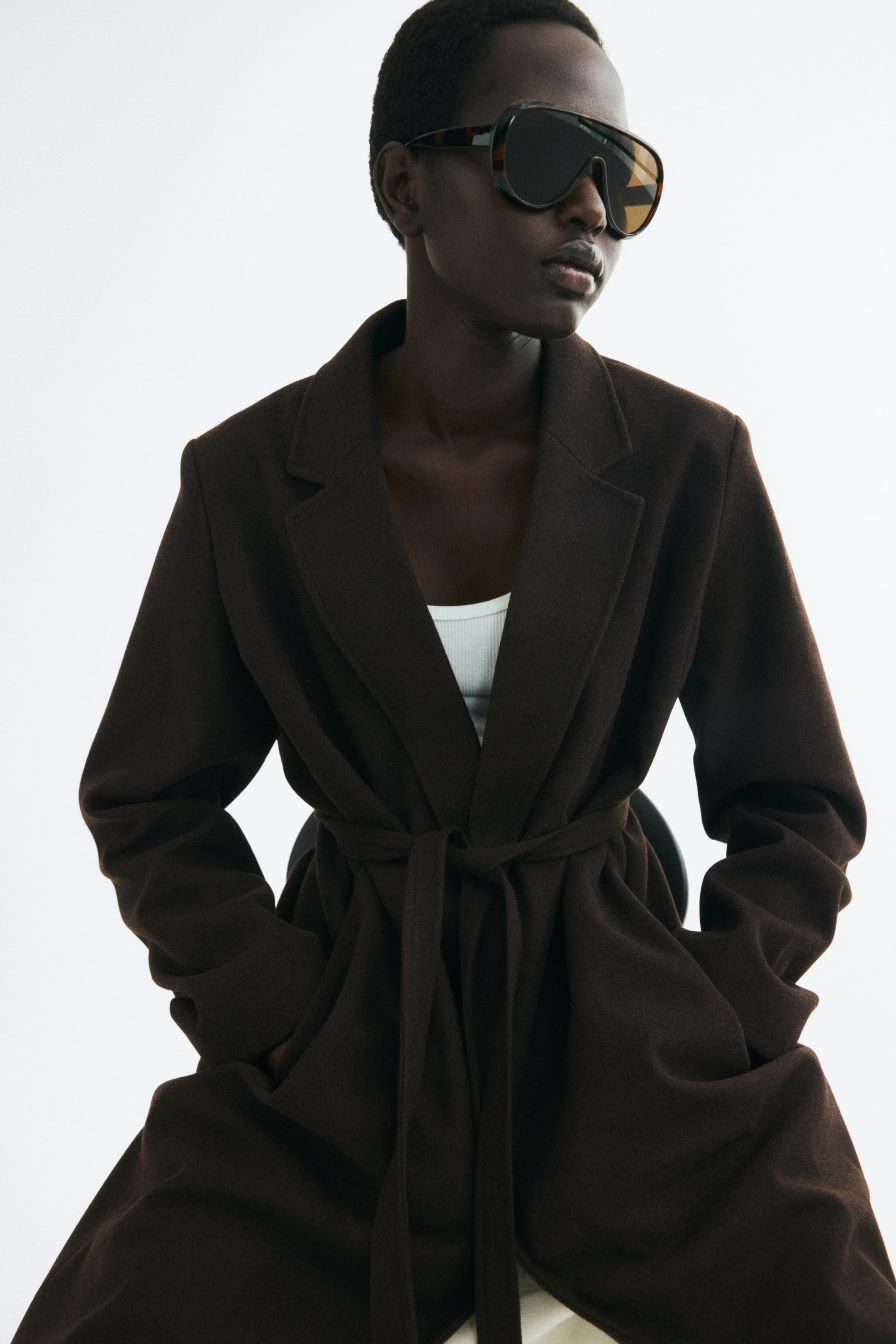 View larger image: A dark brown brushed-weave coat with notch lapels and a tied waist belt is worn by the model. The model is seated with hands in the side pockets, styling the coat over a white ribbed top.