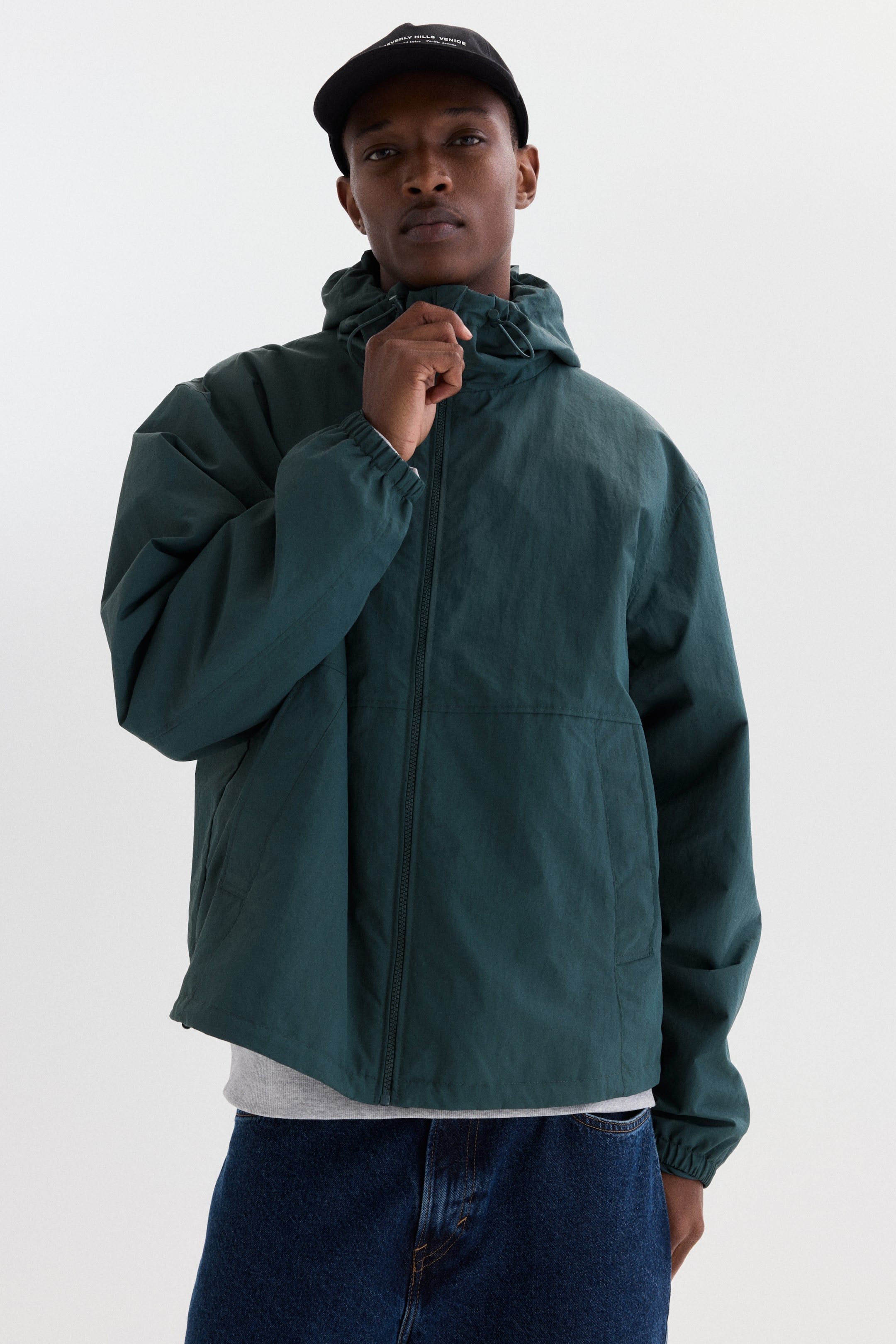 View larger image: A dark green hooded jacket is shown from the front, featuring a full-zip closure and an adjustable drawstring hood. It is designed with long sleeves, elasticated cuffs, and side pockets, appearing with a relaxed fit over a light grey t-shirt.