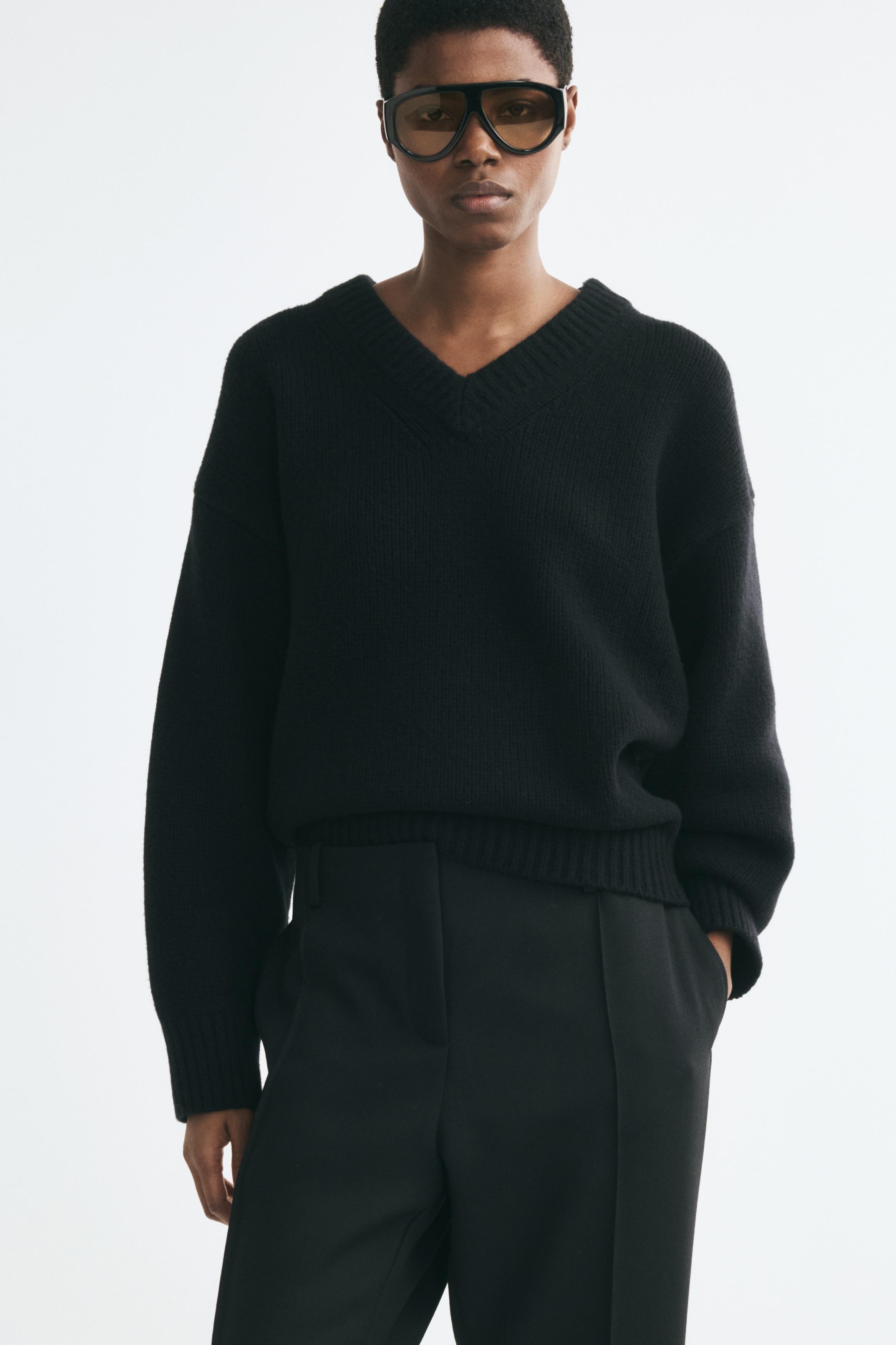 Un pull en laine tricotÃ©e noir, oversize, est prÃ©sentÃ© de face, avec un col en V bordÃ© de cÃ´tes, des Ã©paules tombantes et des manches longues avec poignets cÃ´telÃ©s, ainsi quâun ourlet cÃ´telÃ© assorti.