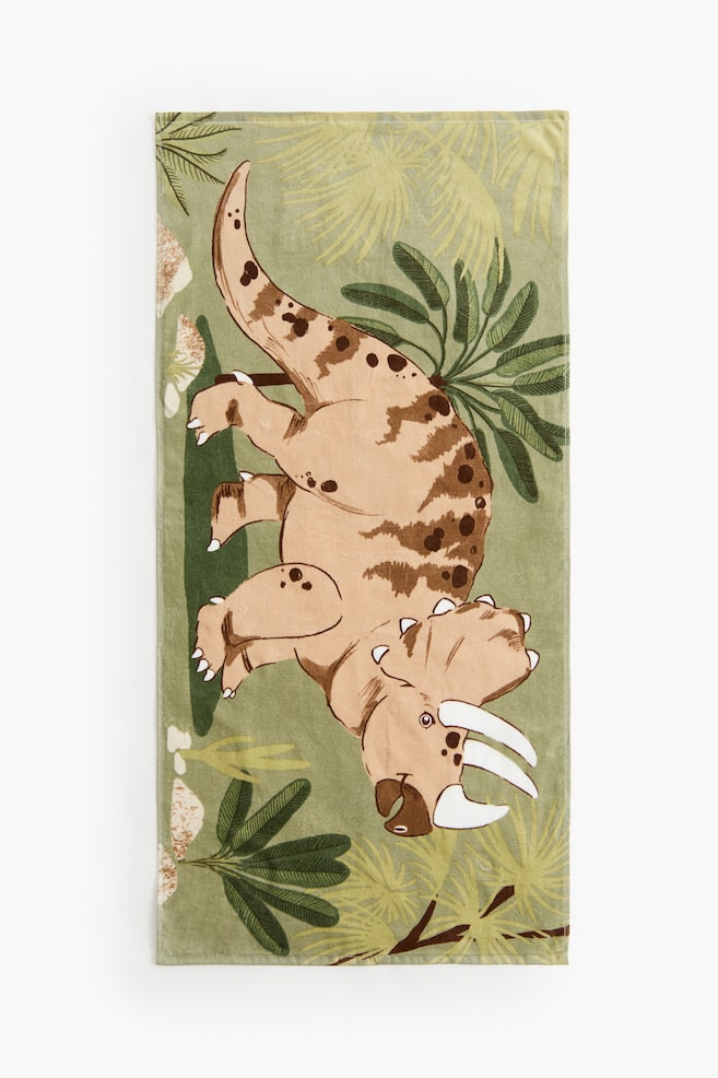 Une serviette de bain rectangulaire affiche son DEVANT, mettant en valeur une texture de velours avec une base vert clair. Elle est ornée d'une grande impression de dinosaure Triceratops marron clair, rehaussée de taches marron foncé et de cornes blanches, entourée de diverses feuilles tropicales vertes. - 1