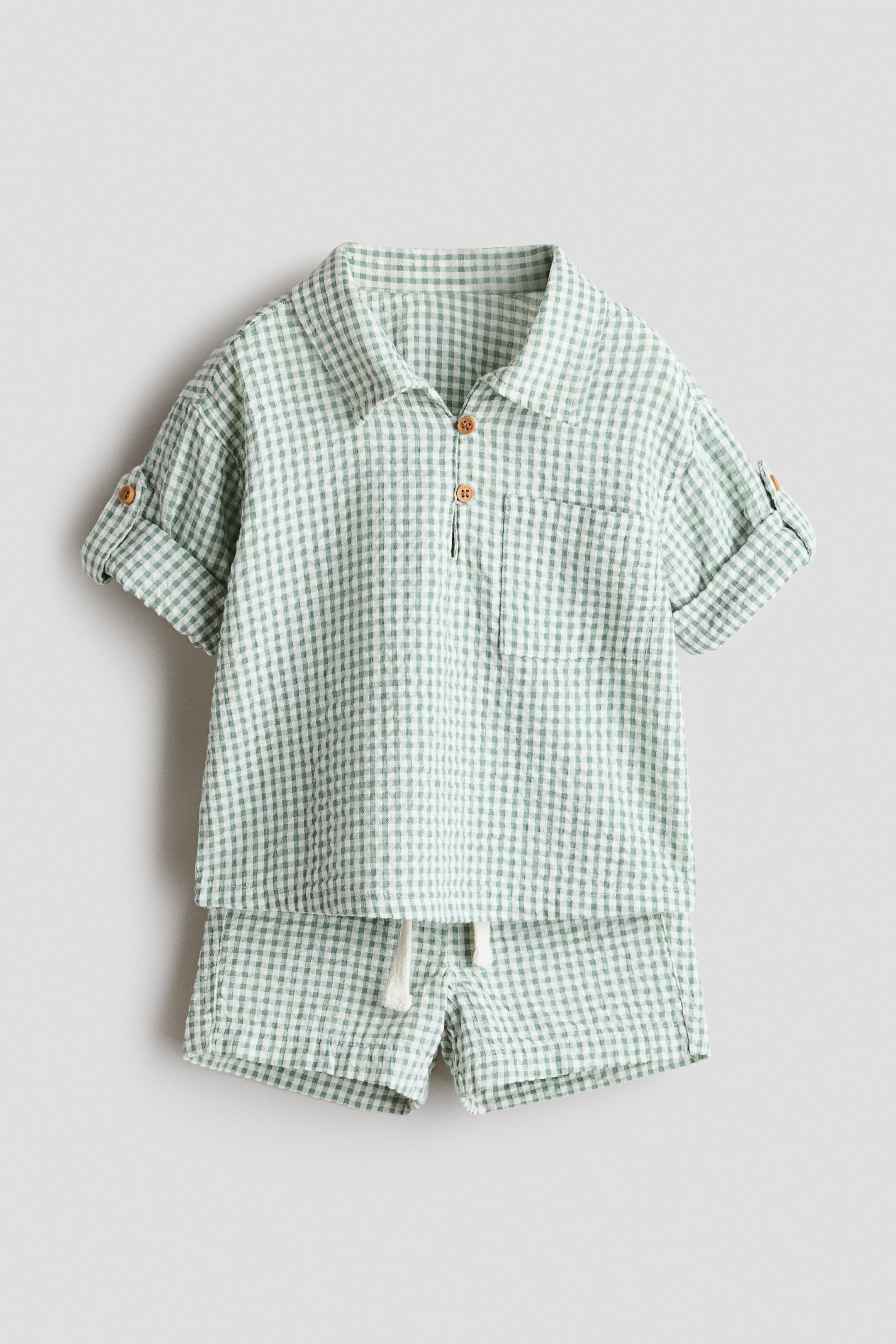 Grotere afbeelding bekijken: Een tweedelig groen en wit geruit outfit wordt gepresenteerd, met een overhemd met kraag en opgerolde korte mouwen die zijn vastgezet met een tab en knoop, een gedeeltelijke knoopsluiting met twee bruine knopen en een enkele borstzak. Bijpassende shorts maken de set compleet, ontworpen met een witte trekkoord in de tailleband.