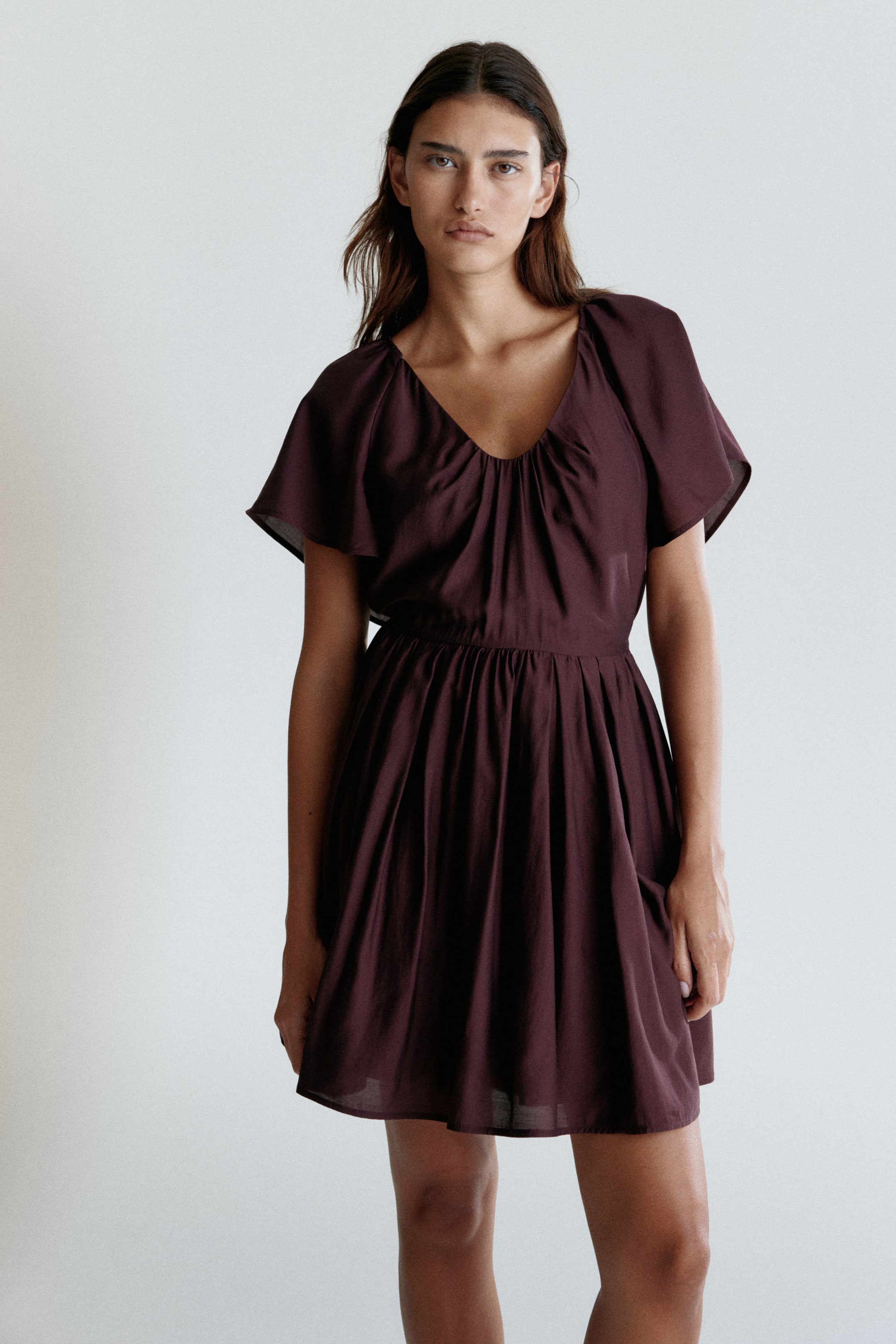 View larger image: A dark maroon dress is facing forward, featuring a V-neckline with central gathers creating a draped bust, and short, wide flutter sleeves. It is cinching at the waist with elasticated gathering and flowing into a short, gathered A-line skirt.