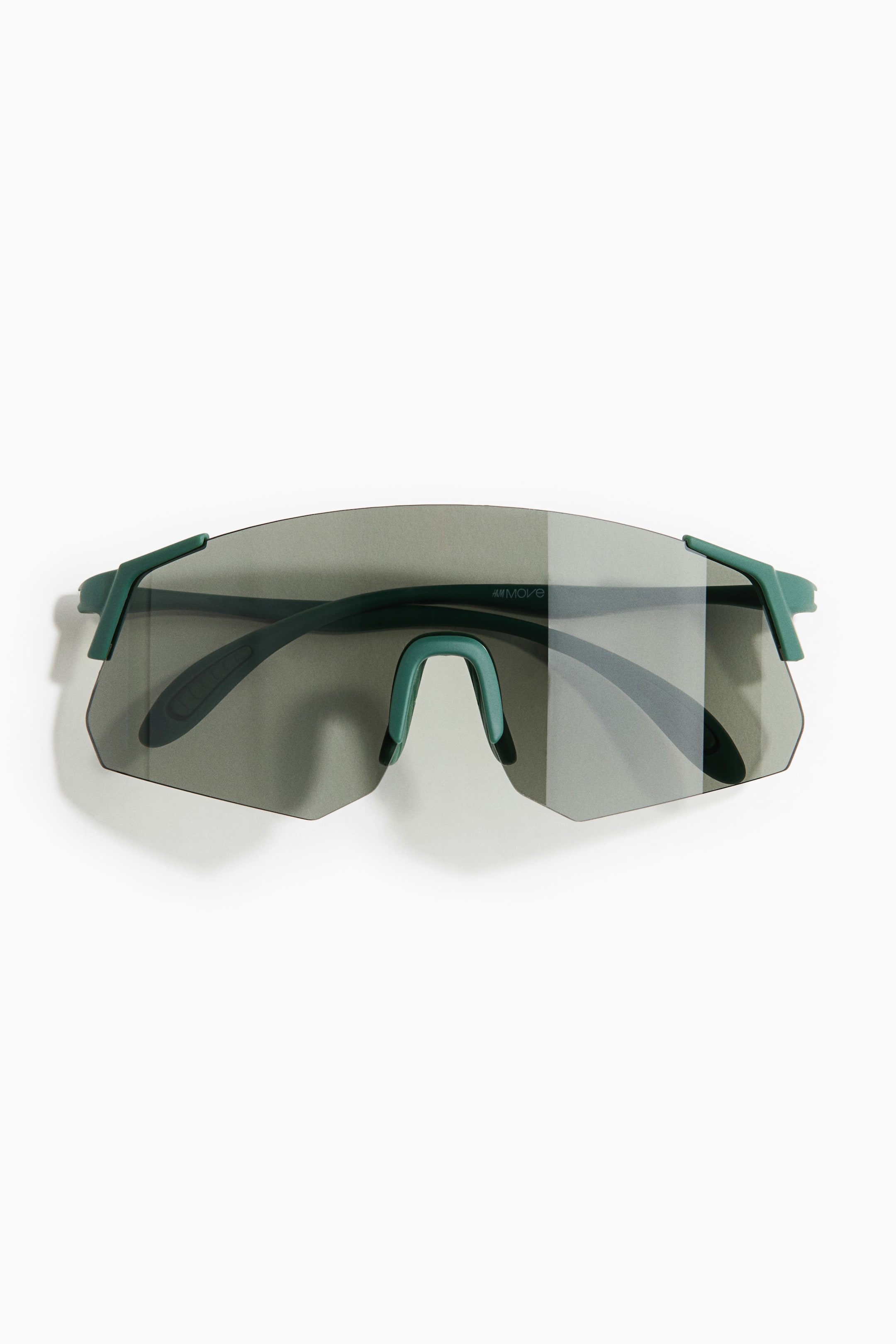 View larger image: Sunglasses ini menampilkan kanta pelindung lebar dengan warna kelabu, serta tangkai berwarna hijau gelap yang memanjang dari sudut atas. Pad hidung berwarna hijau gelap yang terintegrasi terletak di bawah kanta, dan nama jenama "H&M MOVE" dapat dilihat pada bahagian dalam salah satu tangkai.