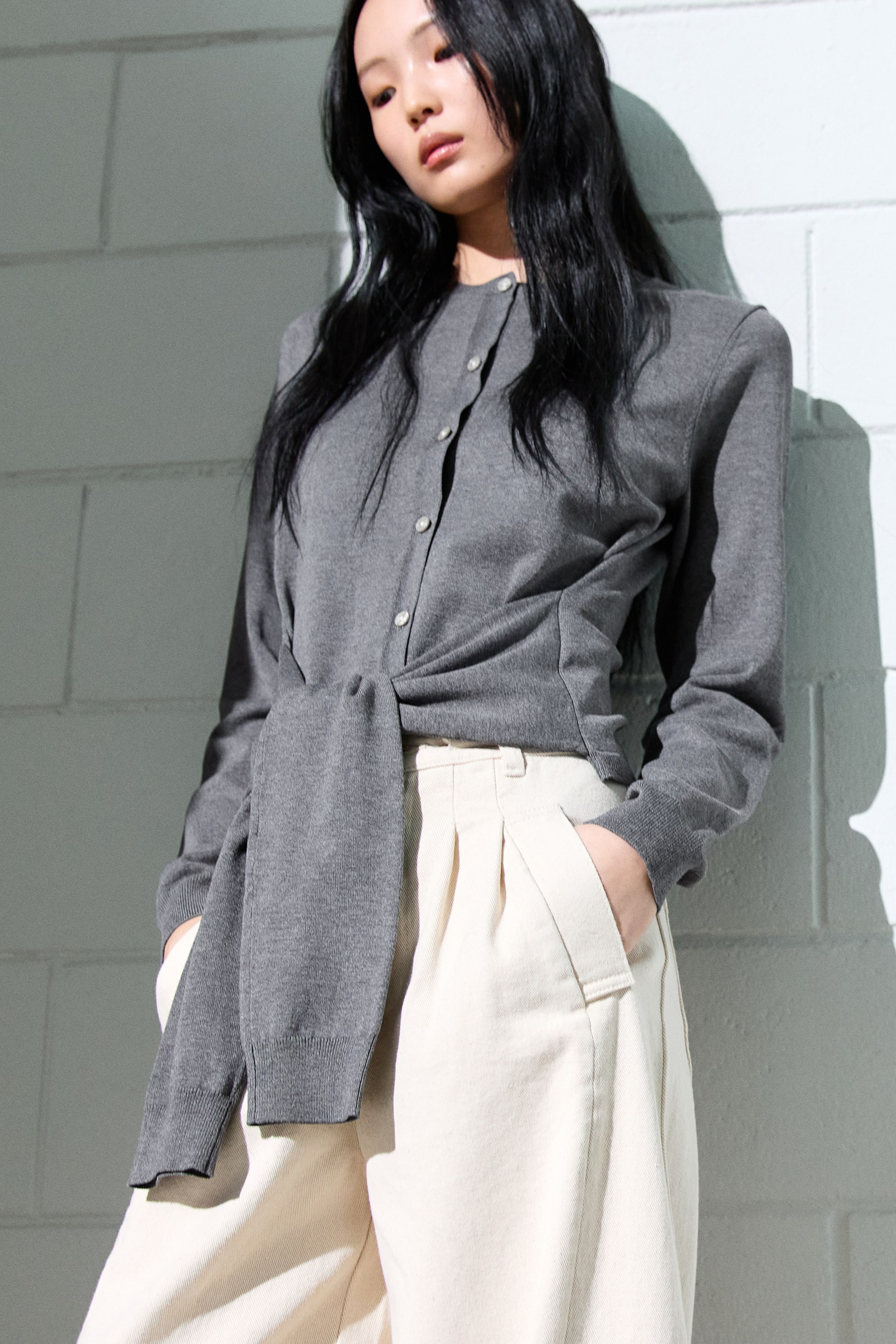 拡大画像を表示: グレーのボタンフロントカーディガンは、クルーネックとリブ付きの長袖が特徴です。ウエストで結ばれたデザインで、フィット感を強調し、クリーム色のプリーツパンツと合わせており、サイドポケットが付いています。