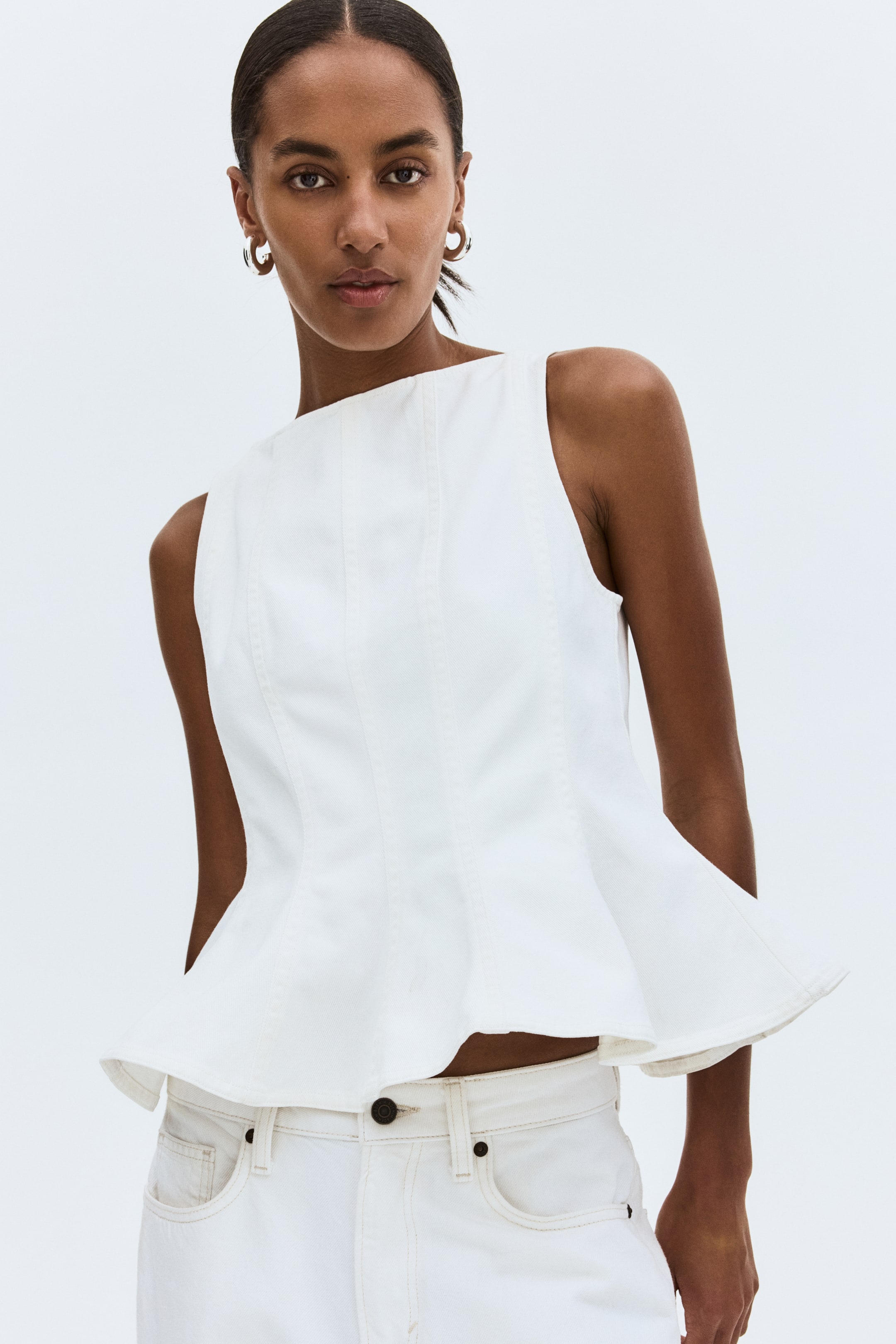 View larger image: A white sleeveless peplum top is displayed, showcasing a high straight neckline and a fitted bodice with vertical seams. Its structured peplum hem flares out, resting above white jeans.