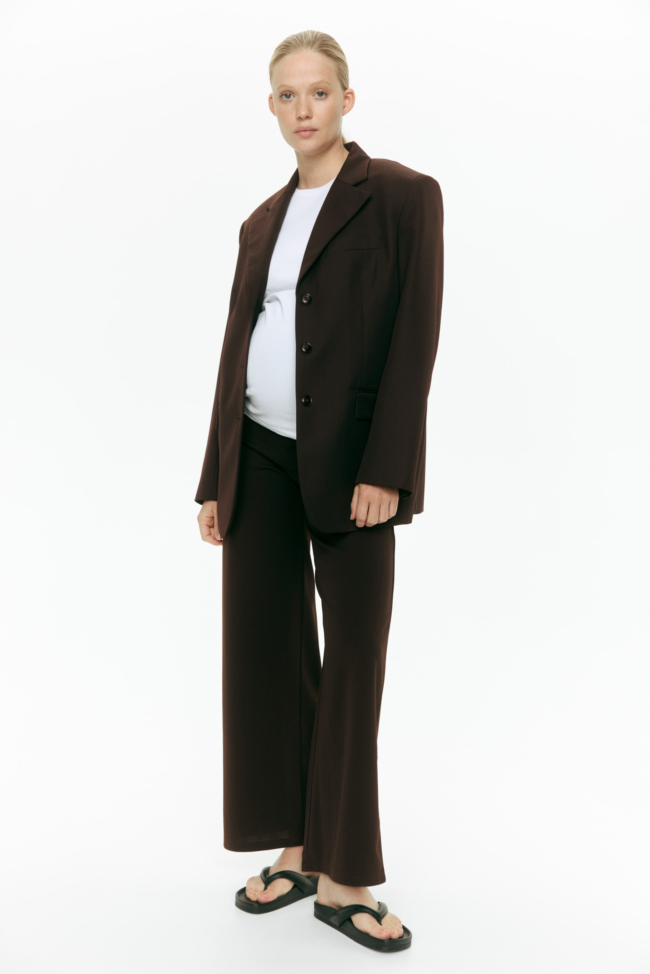 Agrandir l'image: Un blazer marron foncé, doté de revers crantés, de trois boutons à l'avant et de poches à rabat, est porté ouvert sur un haut blanc et s'accorde avec un pantalon large assorti. Le pantalon présente une coupe décontractée sur un pantalon noir, avec l'ensemble complet orienté légèrement vers la droite, mettant en valeur sa structure avant et latérale.