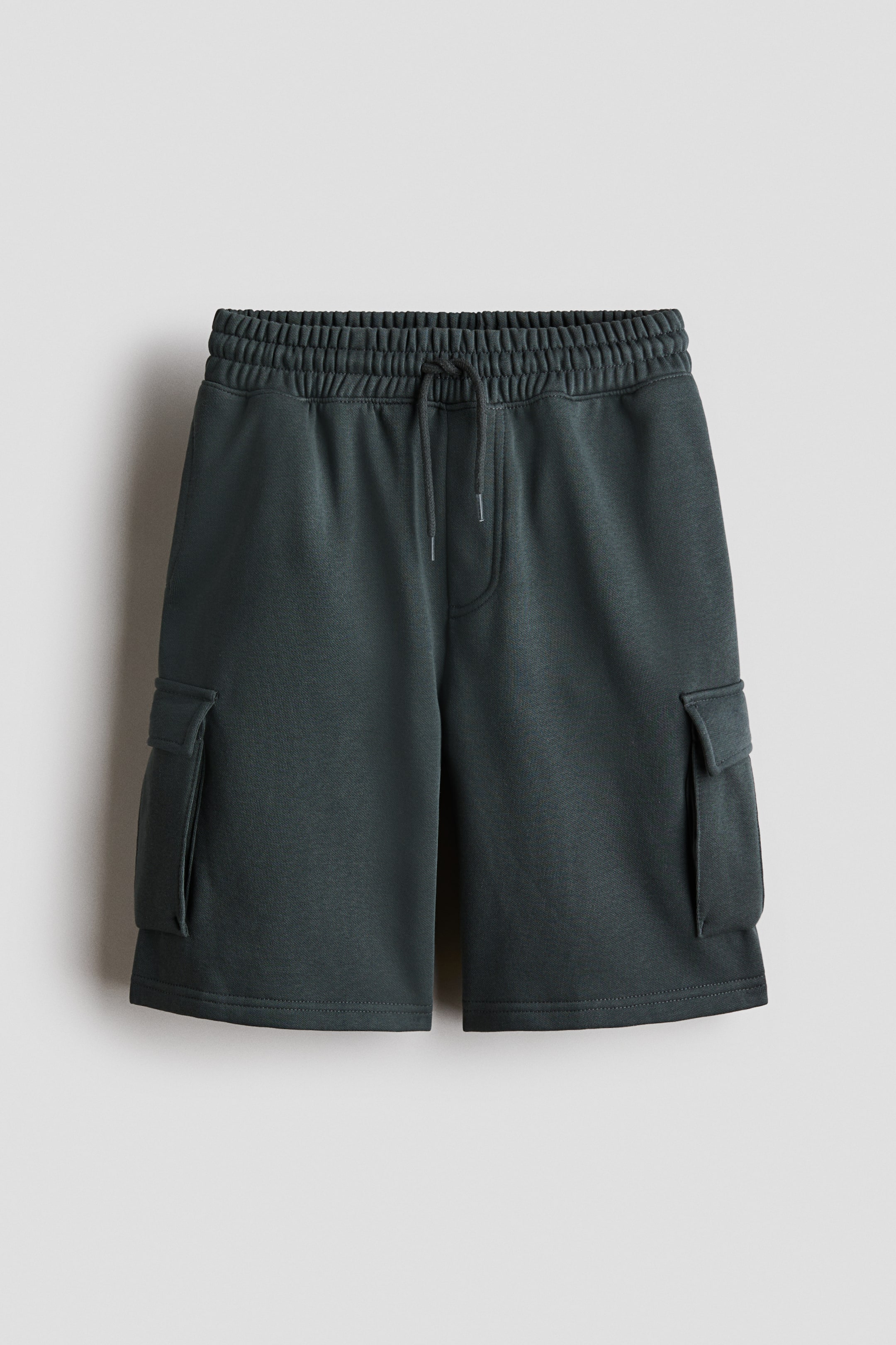 View larger image: Dark gray-green knee-length cargo shorts are showcased in the foreground. They feature a relaxed fit in sweatshirt fabric, an elastic waistband with a drawstring, a faux fly, welt side pockets, and bellows leg pockets with a flap.
