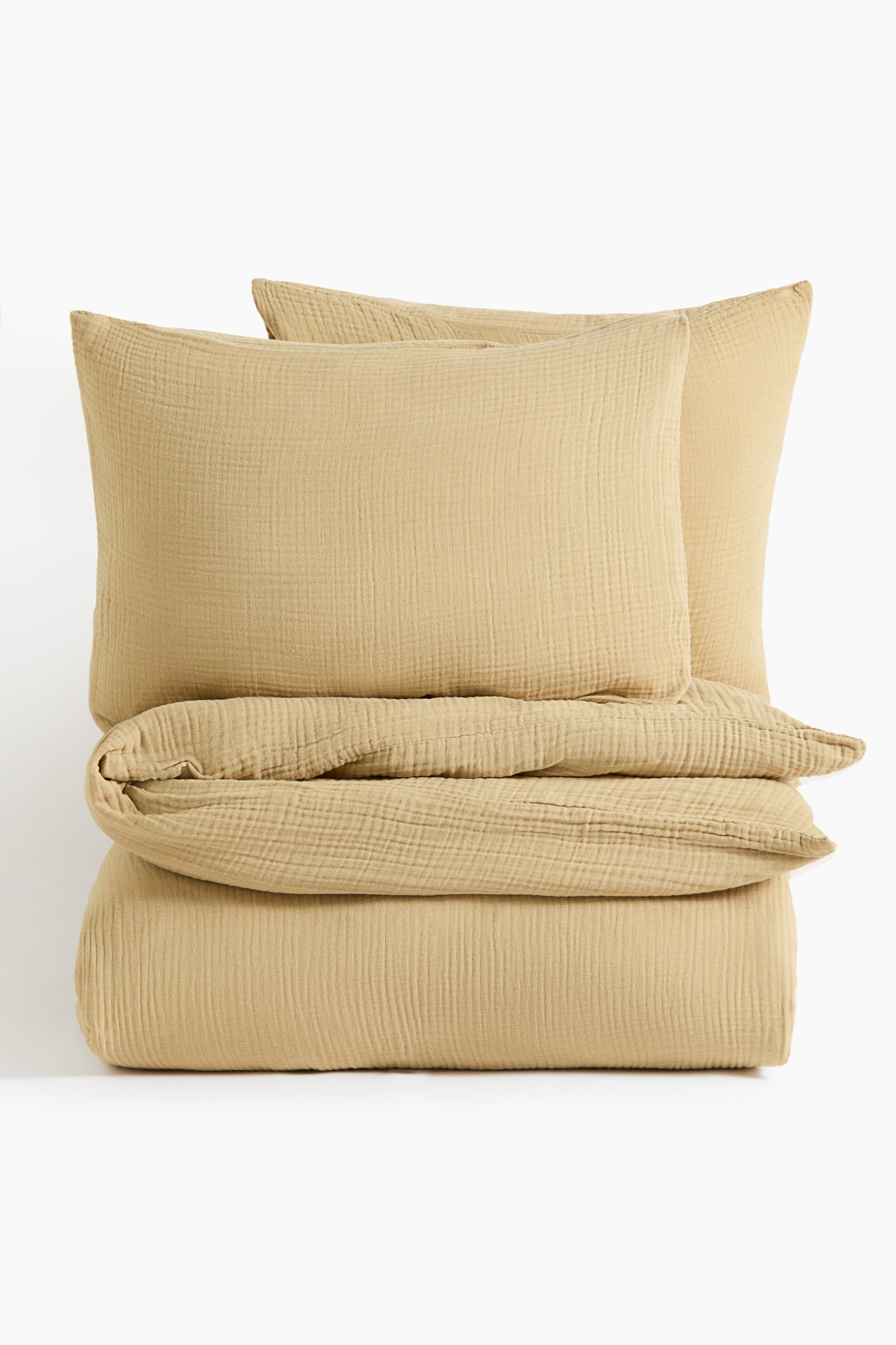A cotton muslin quilt cover set in a muted mustard-yellow hue is shown from the front, featuring a neatly folded quilt cover at the bottom. Two matching pillowcases are resting upright on top of the folded quilt, displaying the crinkled, textured weave of the fabric. - 1