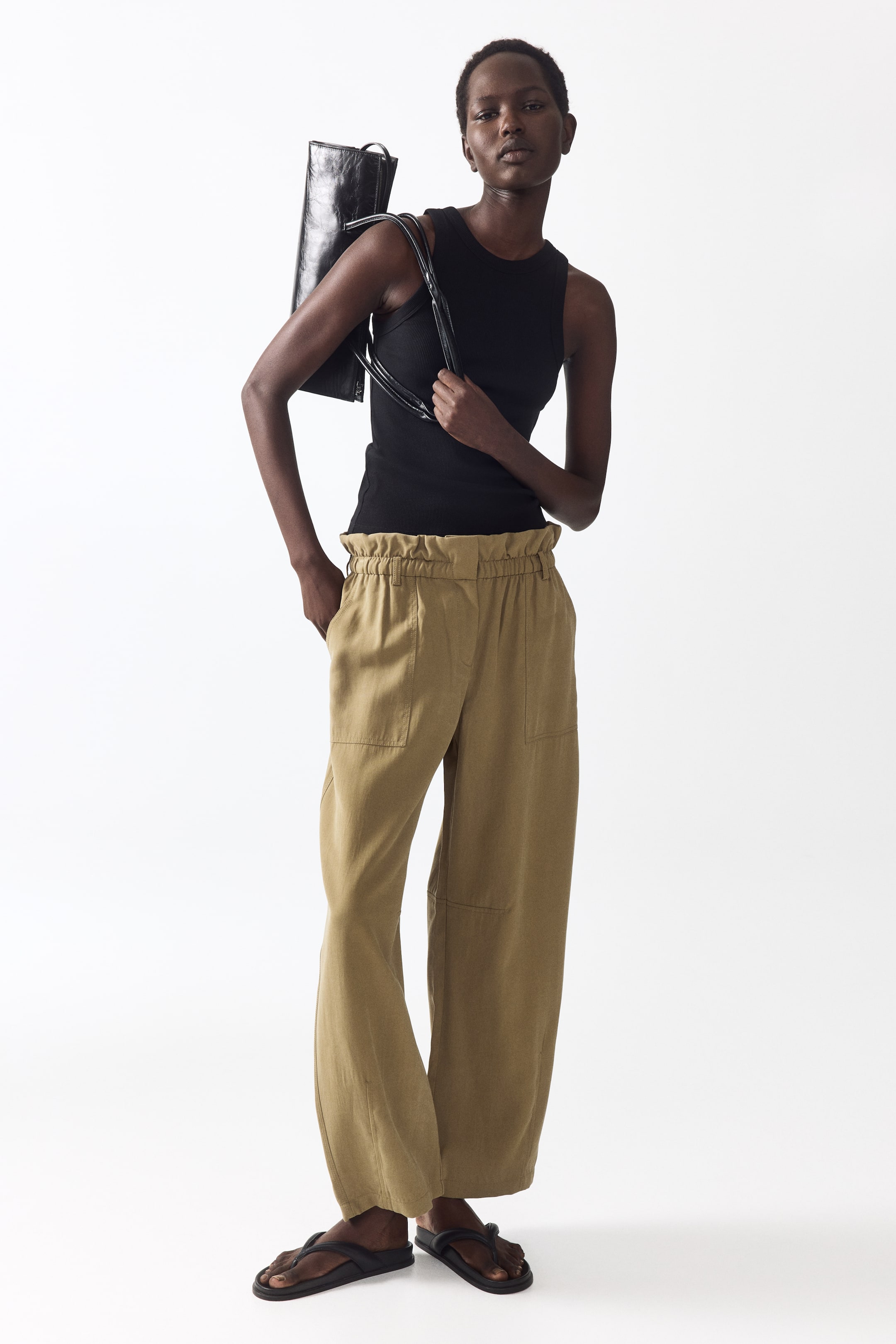 View larger image: Tan wide-leg trousers feature a high, elasticated paper-bag waistband, prominently visible with the top tucked in. They showcase two large rectangular patch pockets on the front and are styled from a slight front-left angle to emphasize the relaxed fit.
