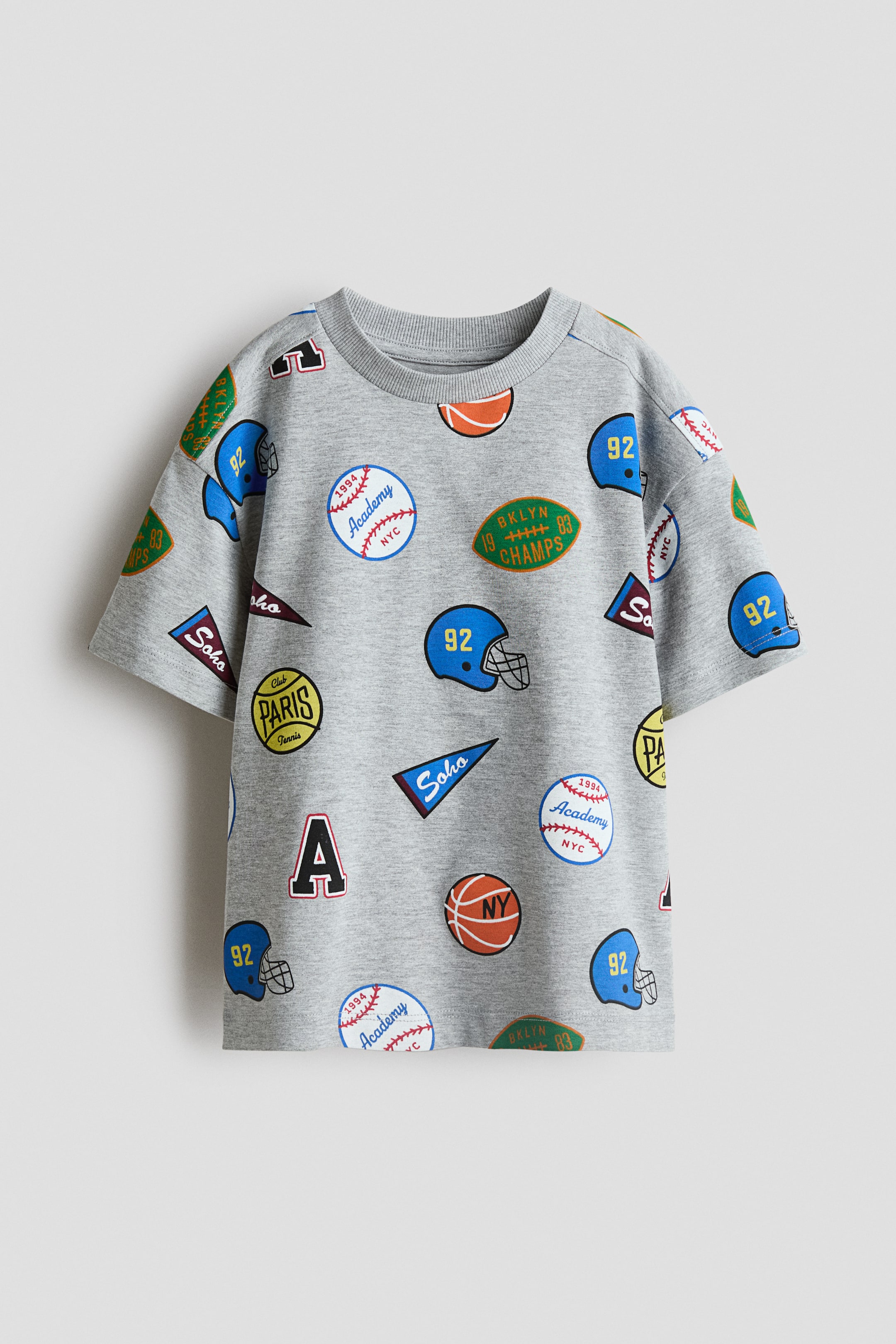 View larger image: A grey T-shirt in soft, printed jersey is presented from the front, featuring a round, rib-trimmed neckline and dropped shoulders. It is patterned with an all-over print of various sports-themed patches, including baseballs, football helmets, and pennants.