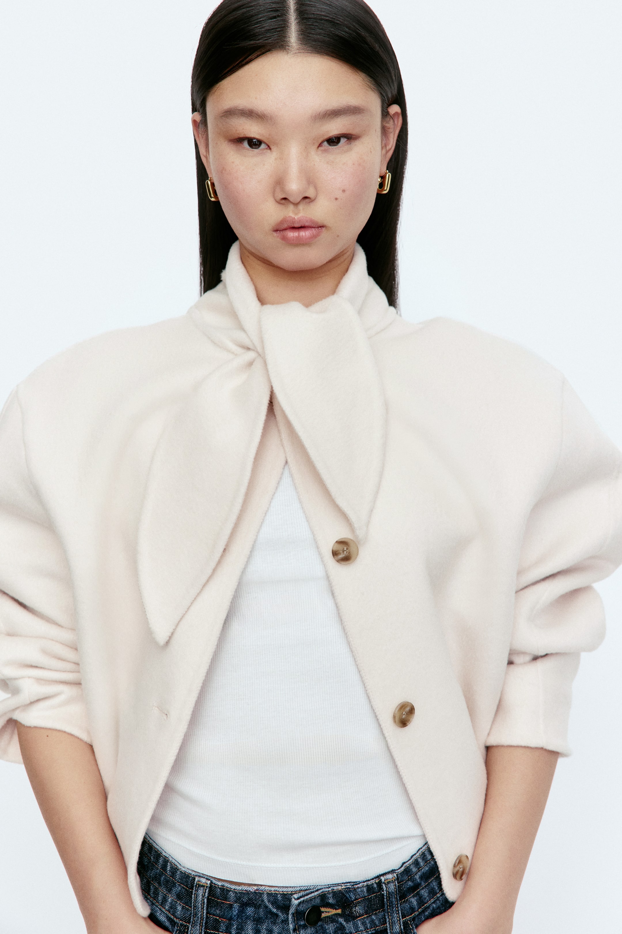 View larger image: A cream jacket featuring an integrated tie-neck collar and a brushed texture, appearing cropped at the waist. Styled open, it reveals a white ribbed top underneath, with light brown button closures visible along the front.