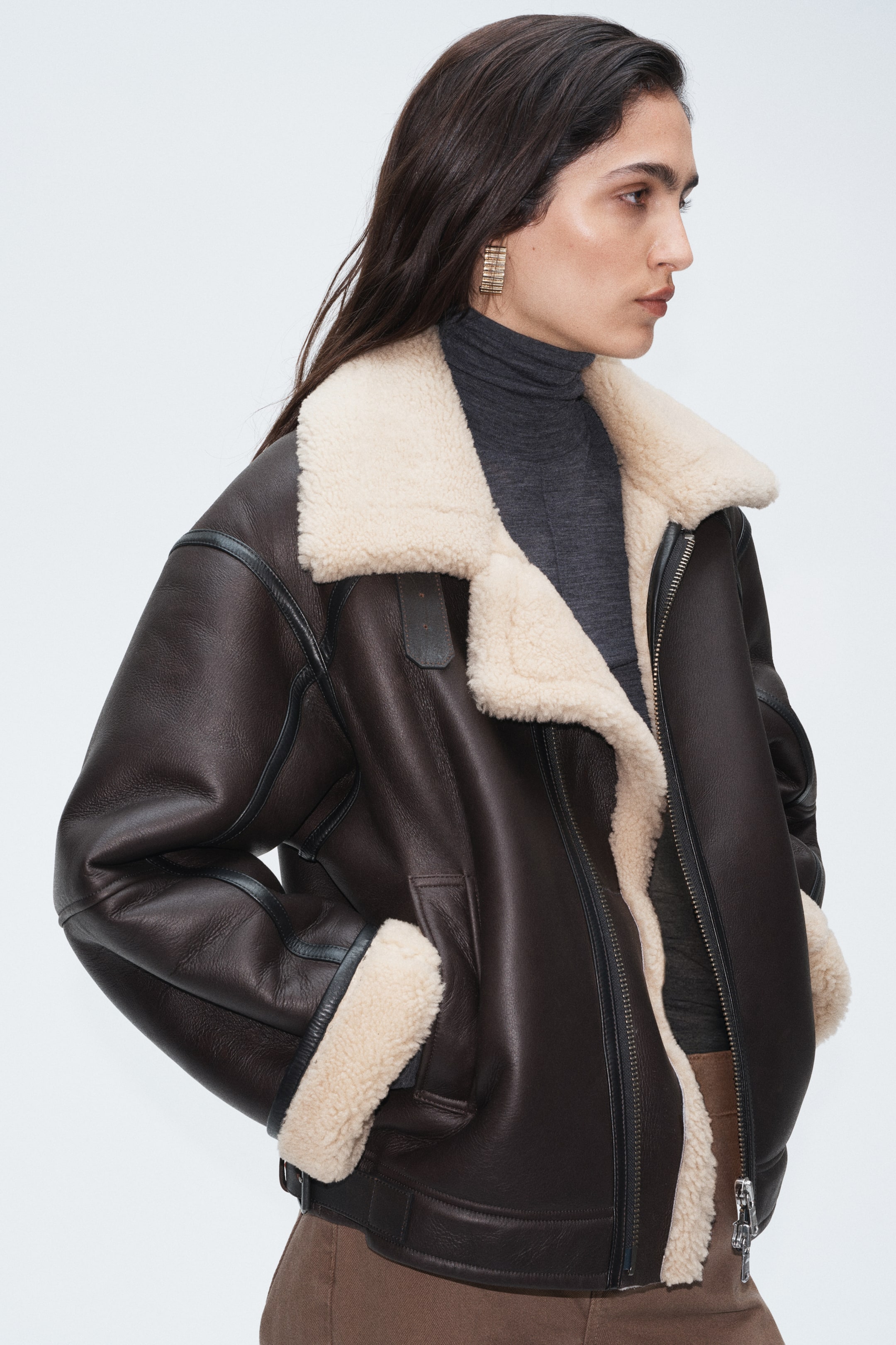 View larger image: A dark brown shearling-lined leather jacket features a wide cream shearling collar, cuffs, and interior lining, with black leather piping. The model is wearing the jacket over a grey turtleneck, with the front zip partially open.
