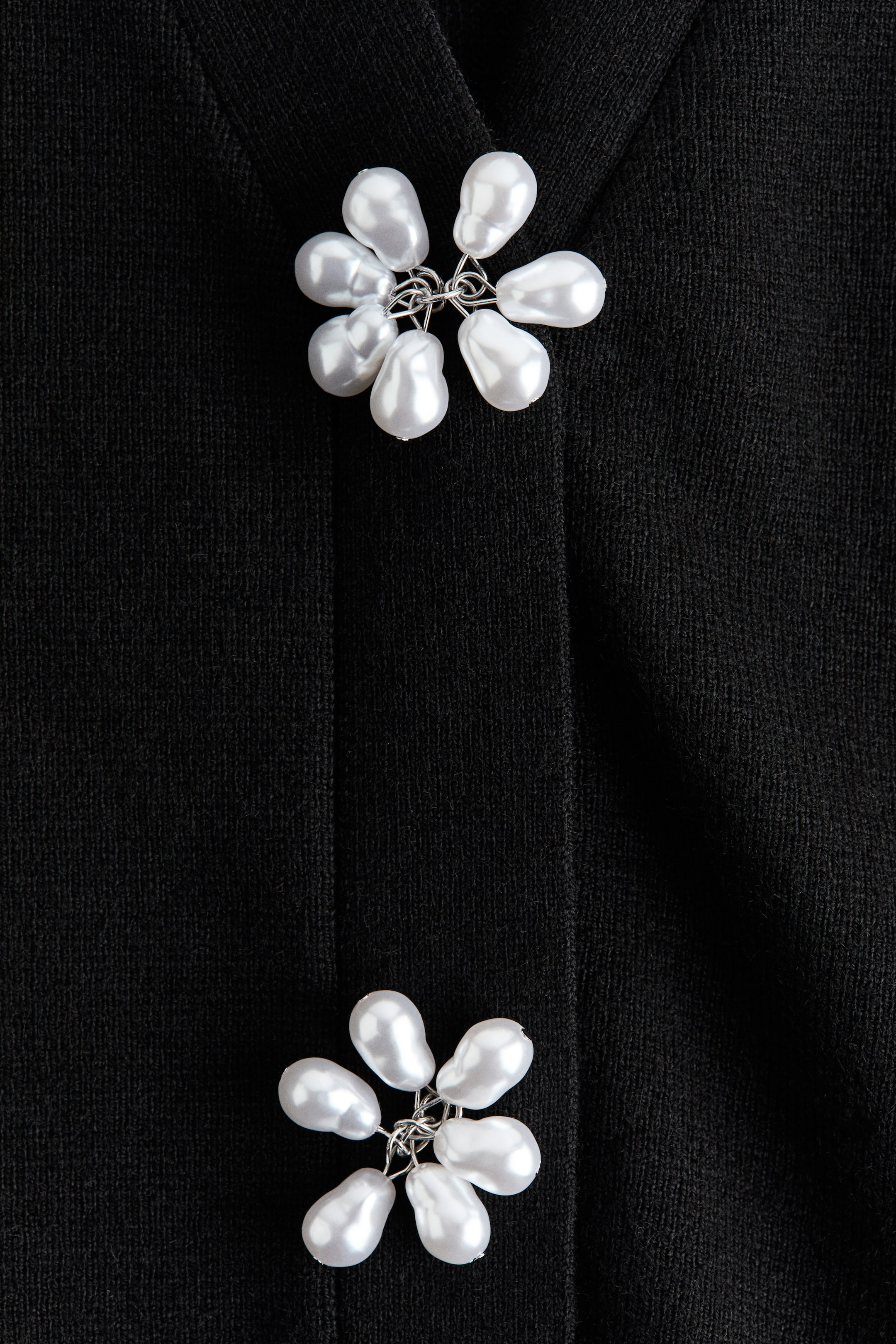 View larger image: A black cardigan is showcasing two decorative flower-shaped buttons down its front placket. Each button is composed of multiple irregular white pearls connected by silver-toned wire, contrasting with the soft knit fabric.