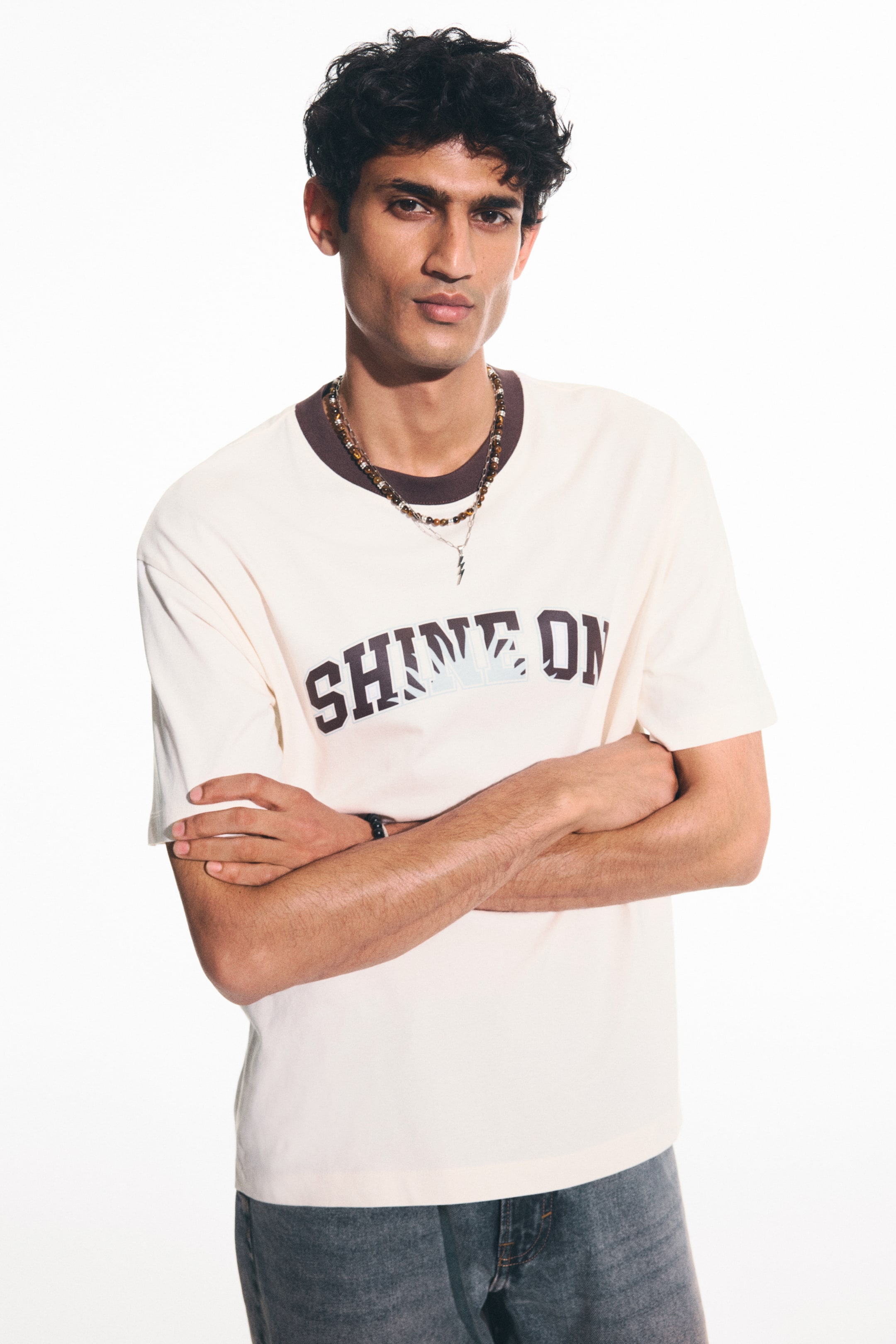View larger image: The cream t-shirt showcases a contrasting brown ribbed crew neck along with short sleeves. It features a dark brown "SHINE ON" graphic print across the chest, incorporating a light blue and white sun design within the lettering.