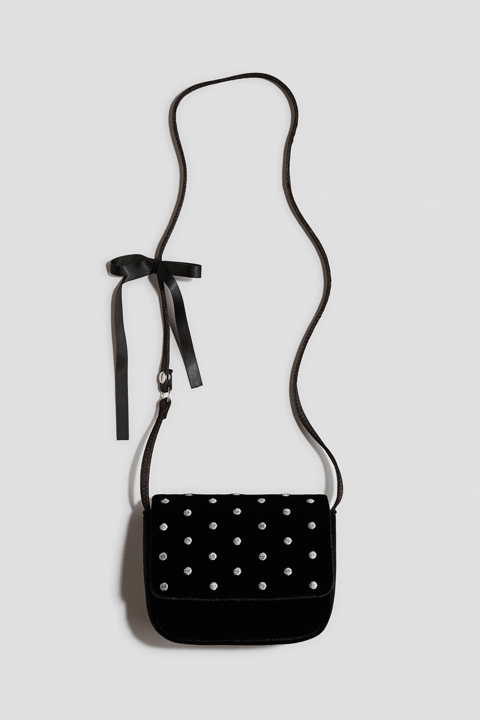 A black velvet crossbody bag is hanging vertically, displaying its front flap embellished with a grid pattern of small, silver-toned rhinestones. The slender black shoulder strap features a black satin ribbon tied into a bow on one side. - 1