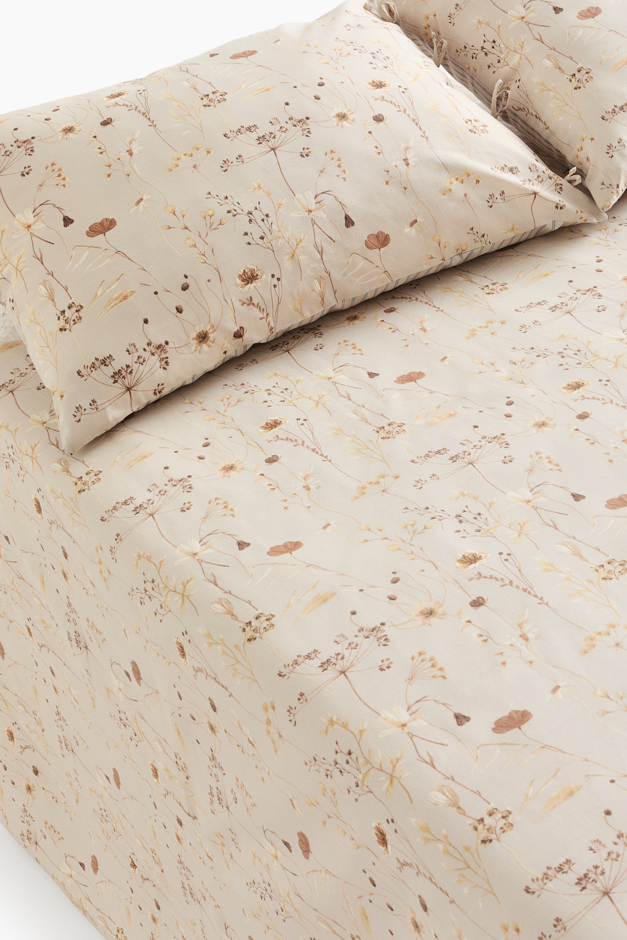 View larger image: A queen bed sheet set featuring a flat sheet and a pillowcase, both showcasing a delicate floral pattern of small botanical motifs in shades of brown, beige, and light yellow on a light neutral background. The pillowcase has ties on one side, and subtle vertical stripes are visible along its edge.