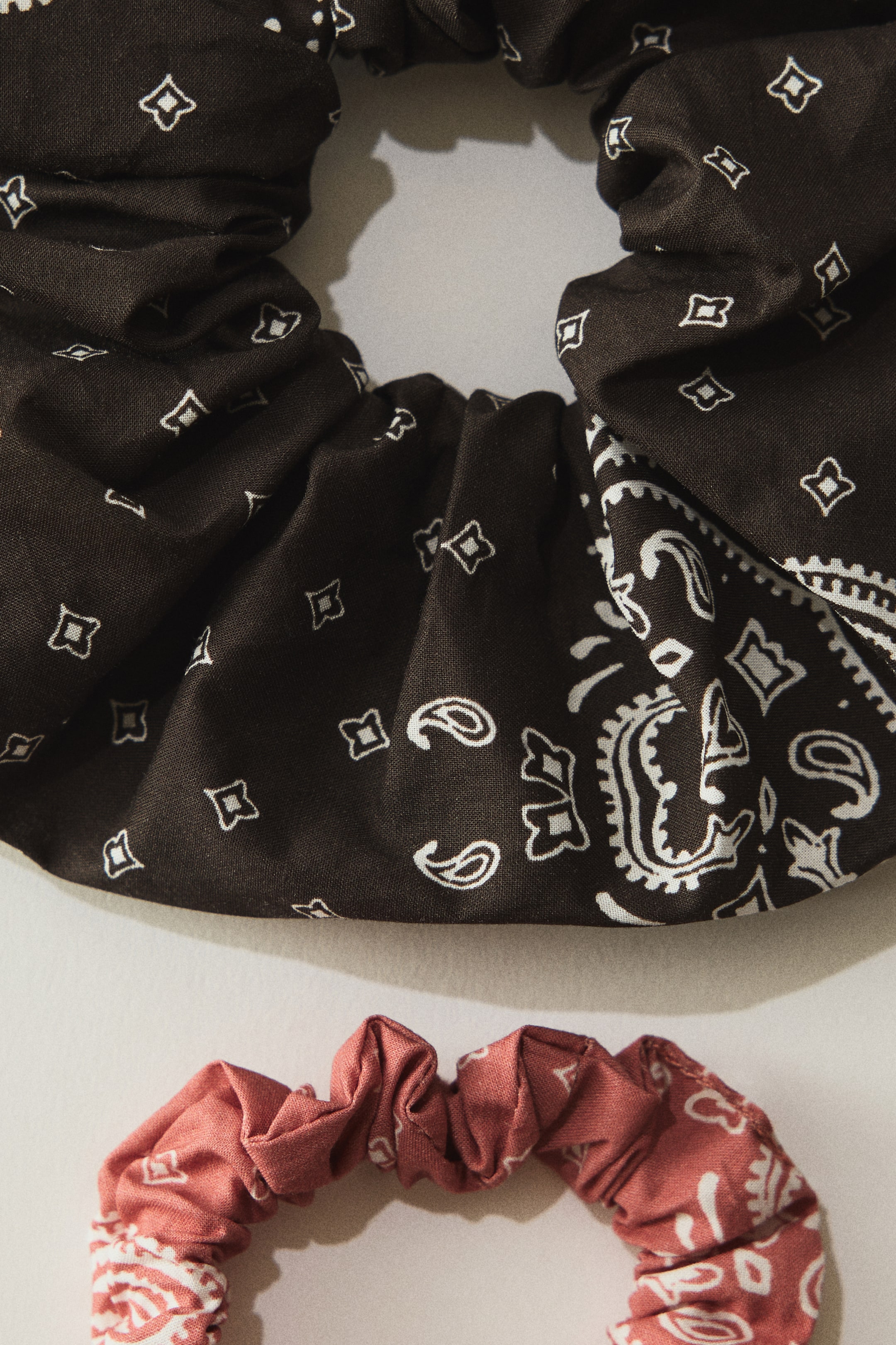 Größeres Bild ansehen: Zwei Scrunchies liegen flach und sind aus der Draufsicht zu sehen. Das obere Scrunchie besteht aus dunkelbraunem Stoff mit einem weißen Bandana‑Muster, das Paisley‑ und Quadratmotiven enthält. Das untere Scrunchie zeigt einen gedämpften roten Stoff mit demselben weißen Bandana‑Muster. Beide weisen eine geraffte Textur auf.
