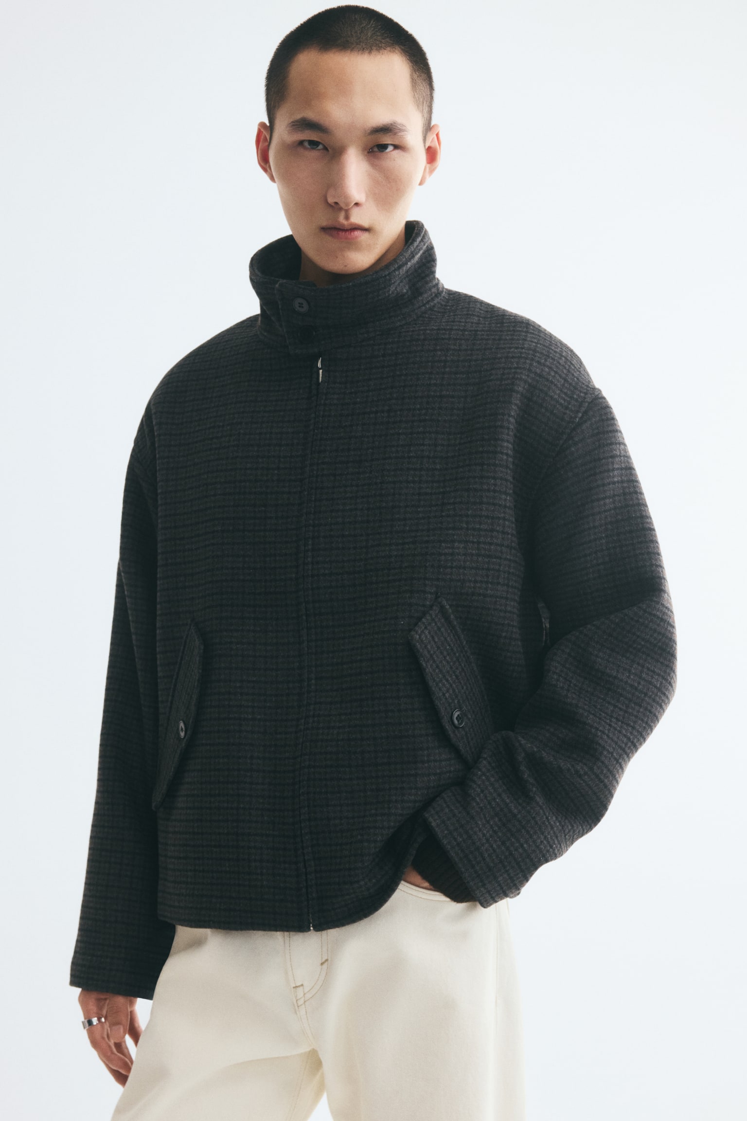 A dark grey micro-patterned jacket is shown from the front, featuring a high funnel neck with dual button closures and a full-length front zip. The jacket is designed with dropped shoulders, long straight sleeves, and two angled front pockets with buttoned flaps. - 1