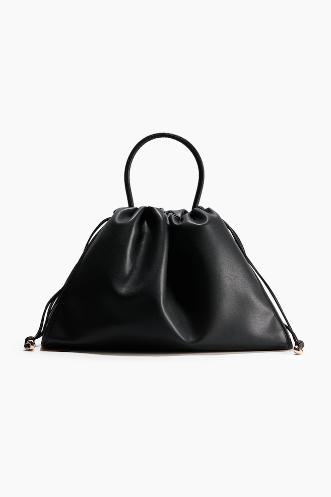 Se muestra de frente un bolso de mano tipo pouch de piel negra, con una silueta holgada y fruncida, y un Ãºnico asa superior estructurada y redondeada. Finos cordones negros de cierre ajustable se extienden desde los laterales, rematados con pequeÃ±as puntas metÃ¡licas en tono dorado. - 1