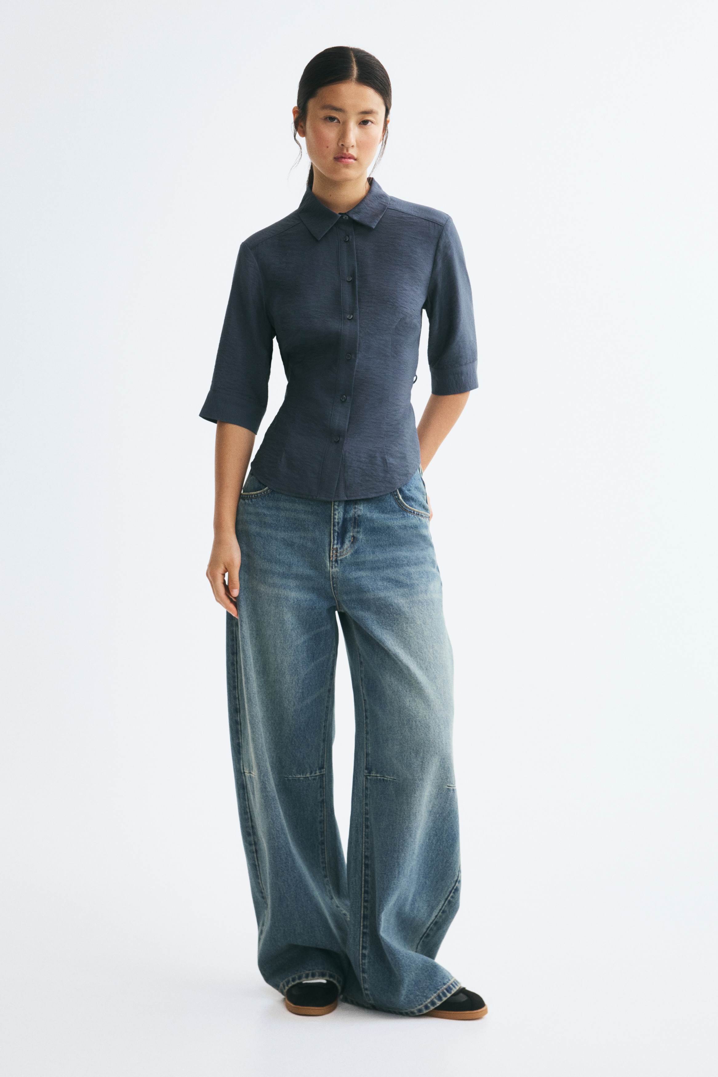 A dark blue button-up shirt is shown from the front, highlighting its textured fabric, point collar, and vertical button placket. It features elbow-length sleeves with wide cuffs and tailored waist seams that create a fitted silhouette.